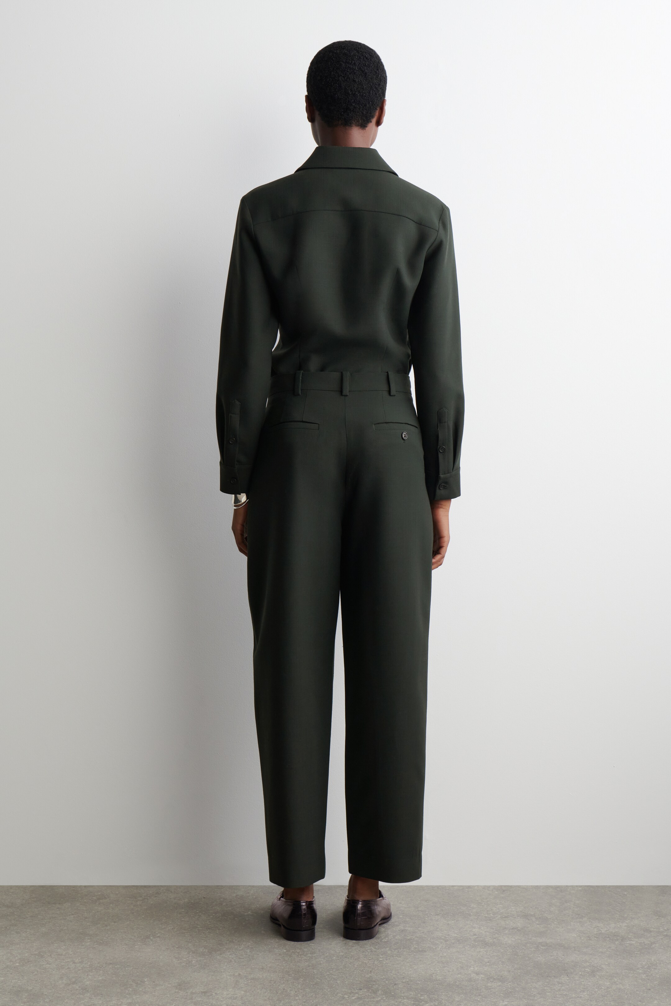 Agrandir l'image: Un tailleur-pantalon vert foncé est exposé de dos. Le haut à manches longues, avec un col classique et un empiècement dos, est rentré dans un pantalon à jambes larges qui présente des passants de ceinture et des poches arrière passepoilées, et est assorti de chaussures plates texturées foncées.