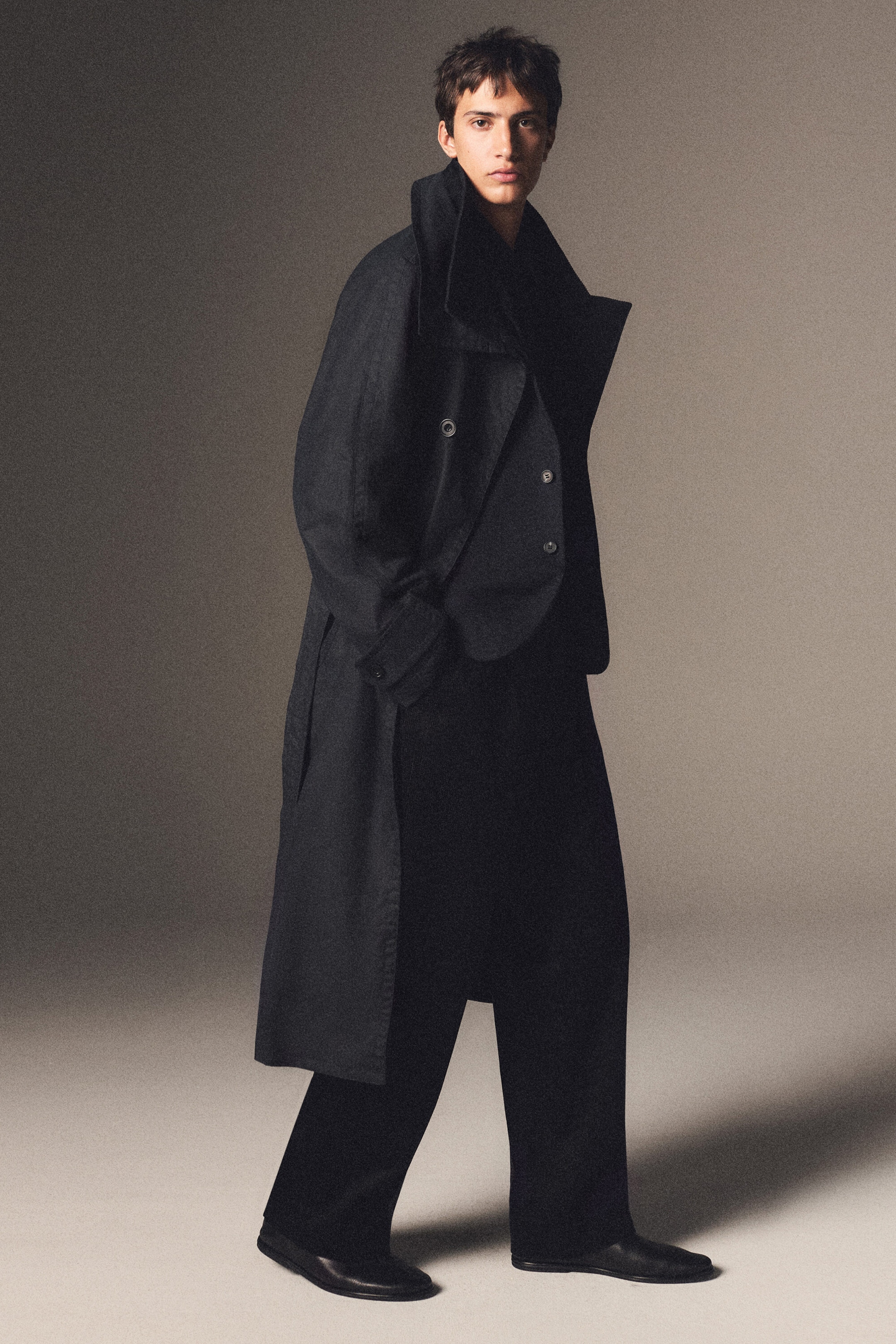A dark waxed-cotton trench coat is shown in a three-quarter view, featuring a long, relaxed silhouette with a high, structured collar and double-breasted button detailing. The coat is styled open over a matching dark blazer and wide-leg trousers, highlighting its fluid drape and layering potential. A dark waxed-cotton trench coat is shown in a three-quarter view, featuring a long, relaxed silhouette with a high, structured collar and double-breasted button detailing. The coat is styled open over a matching dark blazer and wide-leg trousers, highlighting its fluid drape and layering potential.