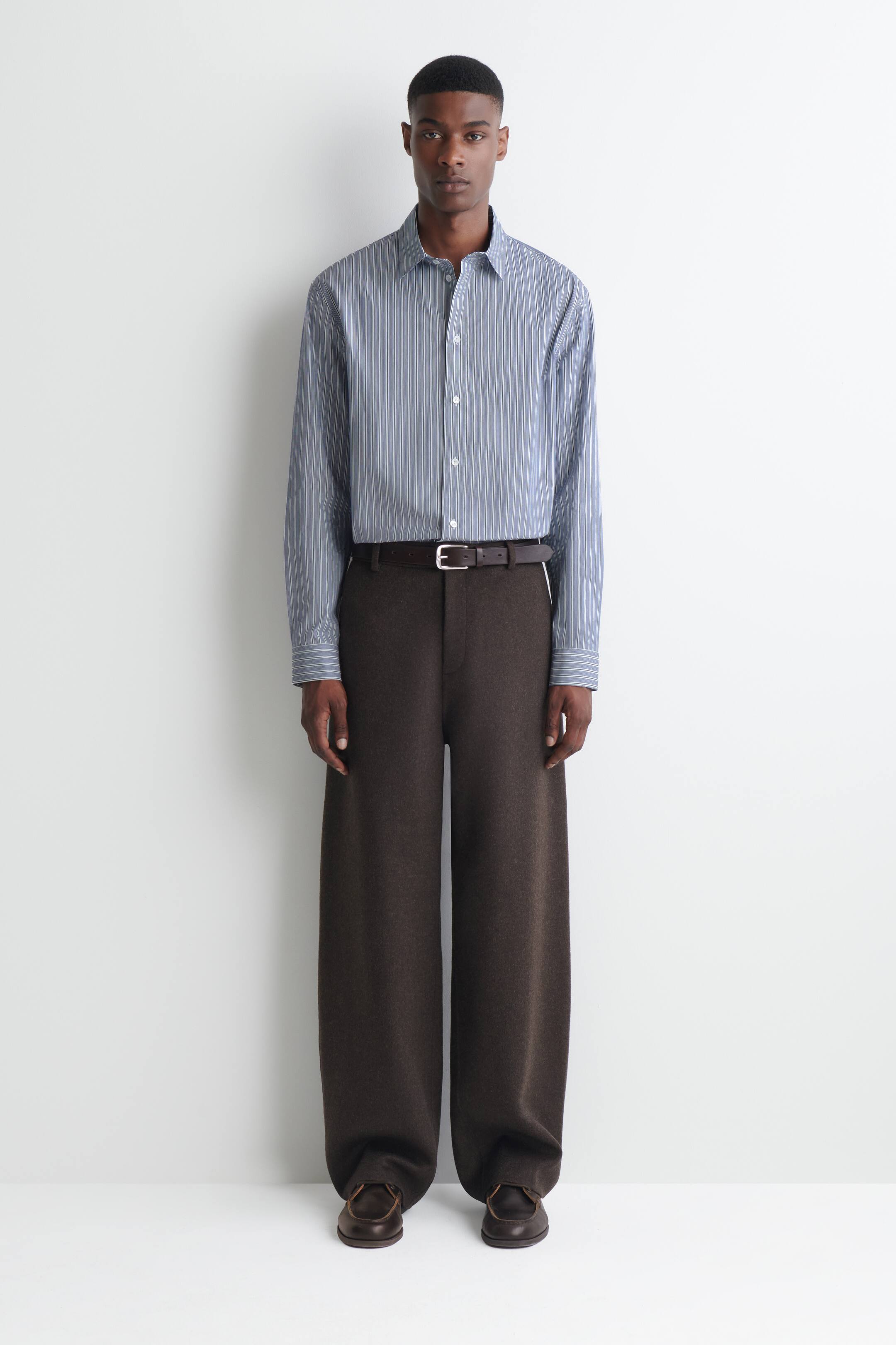 Visualizza immagine più grande: Pantaloni a gamba larga dal taglio rilassato in lana double-face di colore marrone scuro, indossati dalla modella e abbinati a una camicia button-up a righe blu e bianche infilata dentro i pantaloni e a una cintura in pelle marrone scuro. I pantaloni presentano una gamba dritta e ampia che cade sopra scarpe in pelle marrone scuro.