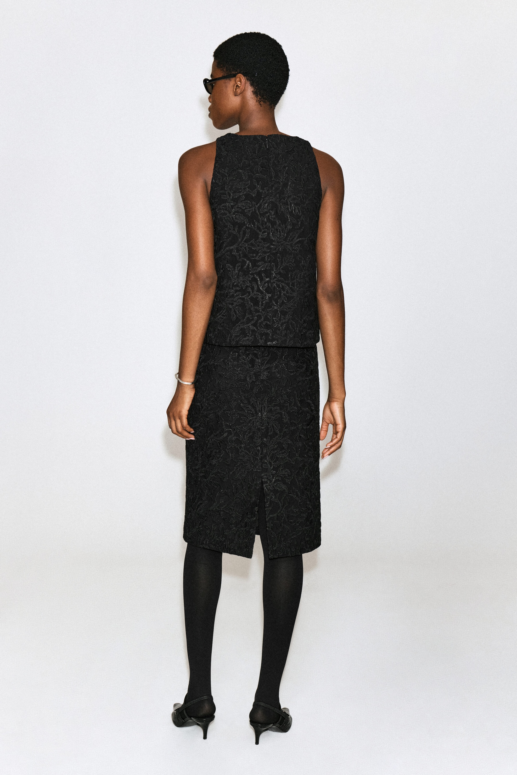 A black lace midi skirt featuring a textured floral pattern and a rear slit is worn by the model, who is facing away from the camera. The skirt is styled with a matching sleeveless lace top, black tights and black heels. A black lace midi skirt featuring a textured floral pattern and a rear slit is worn by the model, who is facing away from the camera. The skirt is styled with a matching sleeveless lace top, black tights and black heels.