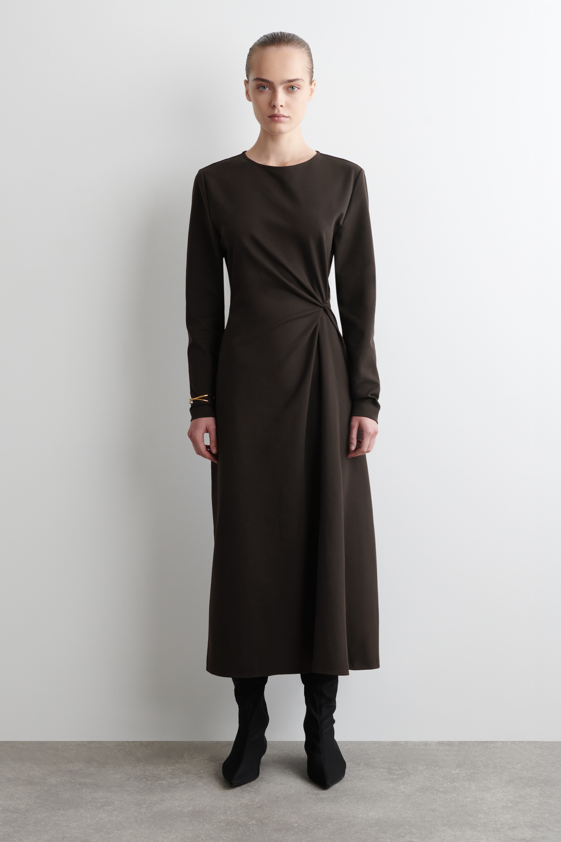 Agrandir l'image: Une robe de ton espresso avec des manches longues, un col rond et un détail torsadé à la taille gauche est portée par un mannequin. La robe a un ourlet légèrement évasé de longueur midi, et le mannequin l'associe à des bottes noires.