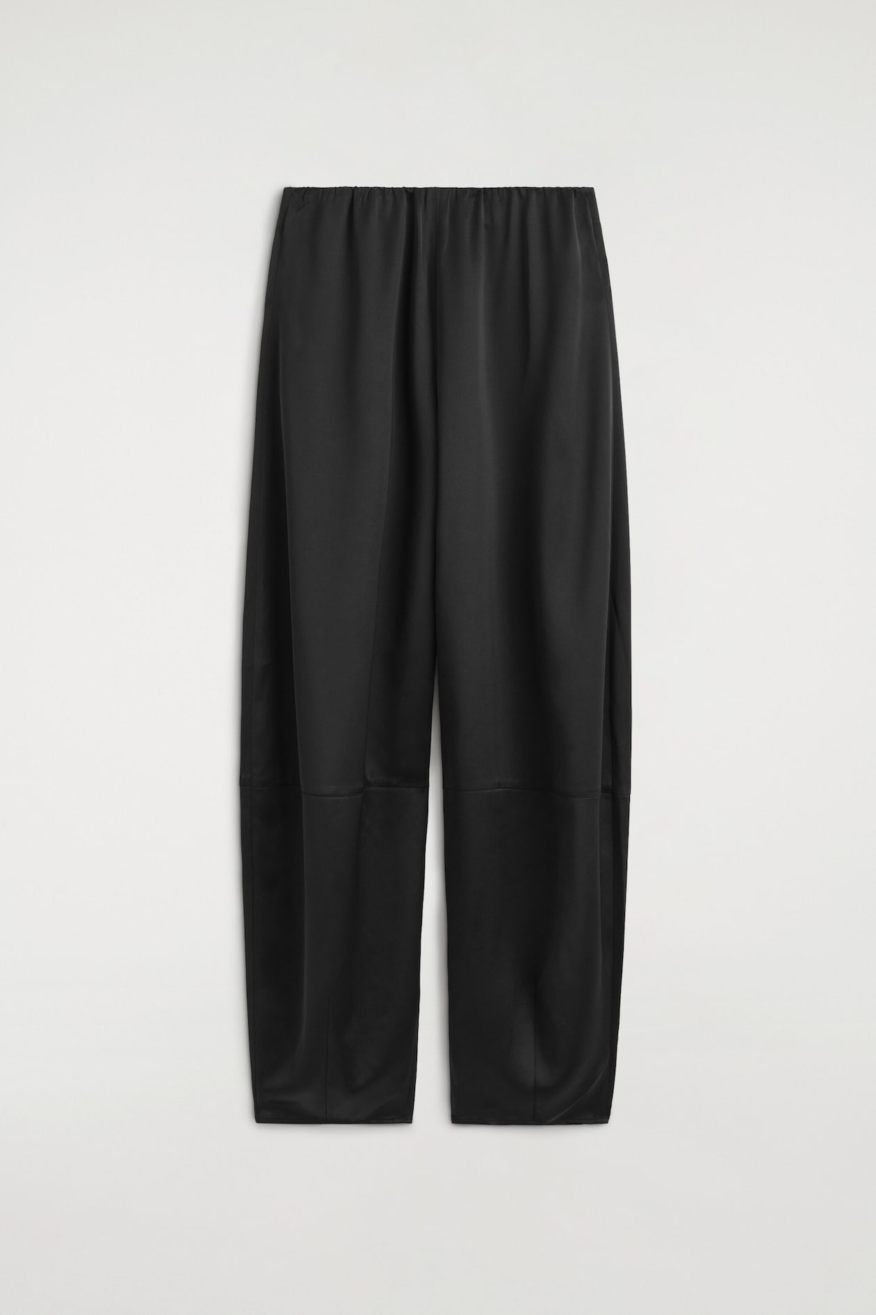 A front view of black satin trousers displaying a relaxed barrel-leg silhouette with a horizontal seam across the knees. The trousers feature a gathered elasticized waistband and a subtly lustrous finish.