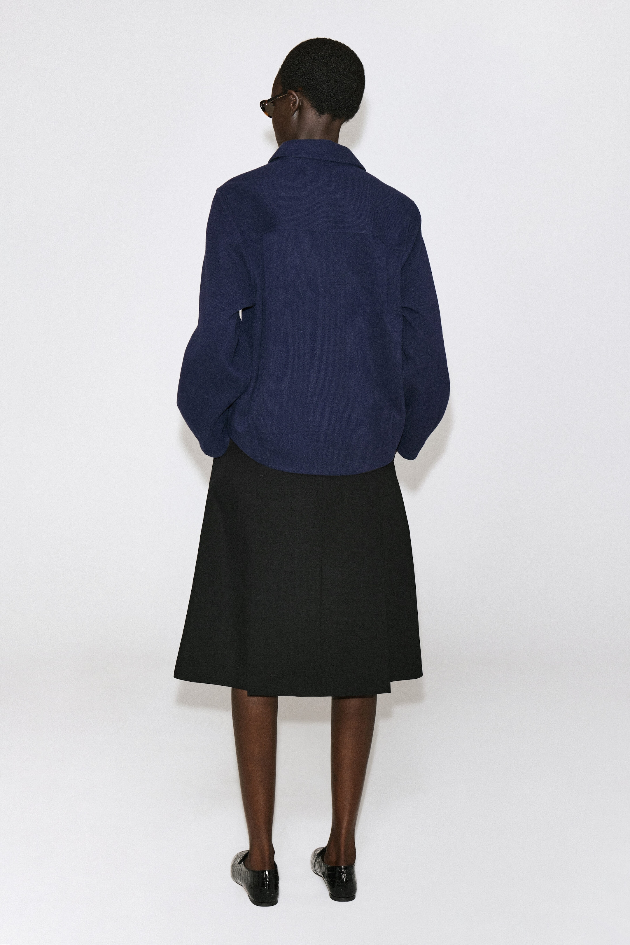 The navy blue corduroy jersey shirt is shown from the back, highlighting its relaxed fit, soft texture, and classic collar. It features a horizontal back yoke and a slightly curved hem, draping loosely over a black knee-length skirt. The navy blue corduroy jersey shirt is shown from the back, highlighting its relaxed fit, soft texture, and classic collar. It features a horizontal back yoke and a slightly curved hem, draping loosely over a black knee-length skirt.