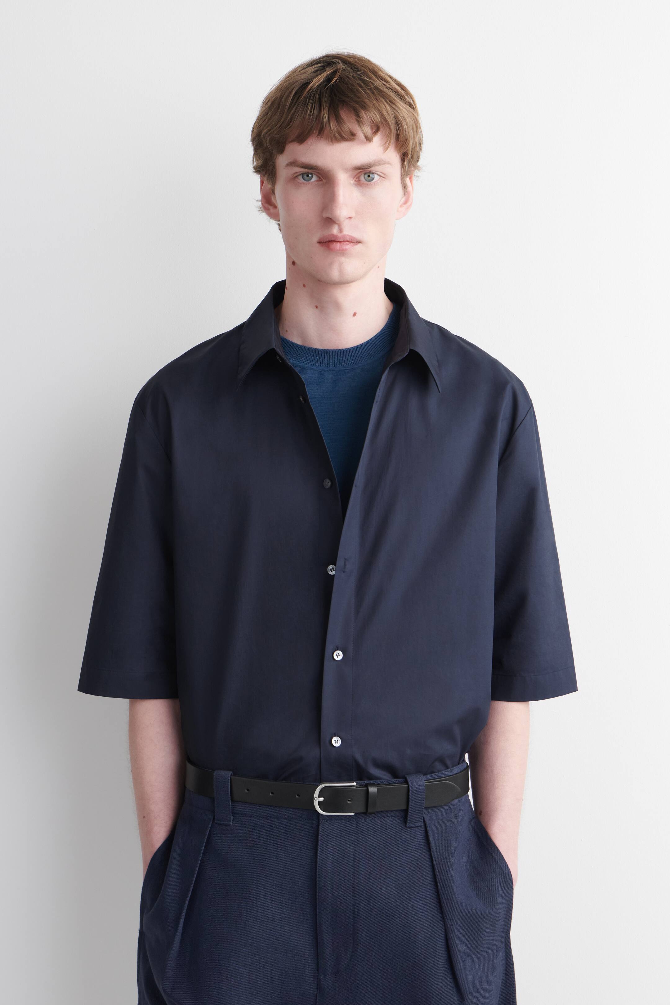 Agrandir l'image: Une chemise bleu marine à manches courtes et coupe décontractée, avec un col pointu et une fermeture à boutons sur le devant, est portée par le mannequin, superposée sur un sous-pull bleu à col rond.