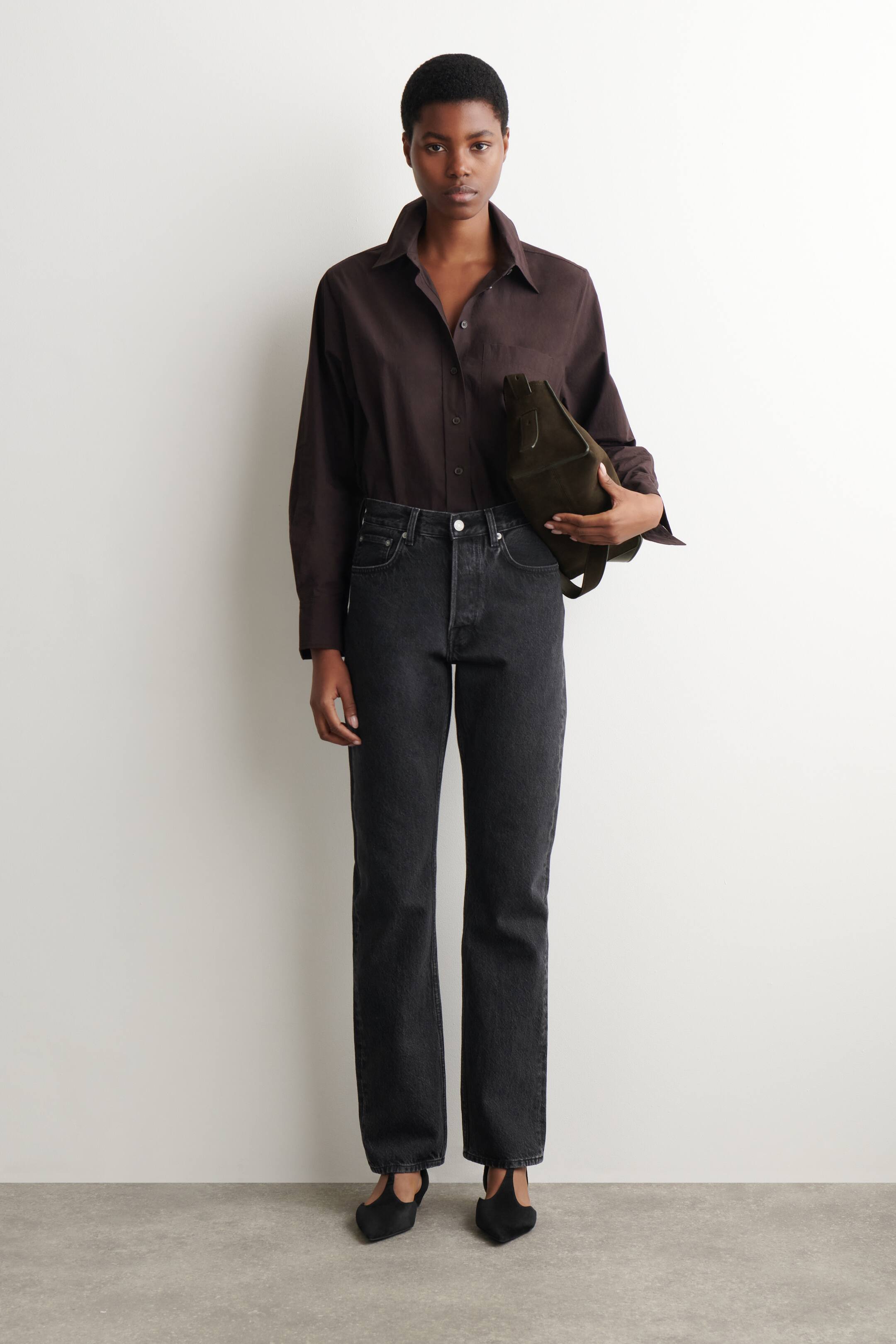 Agrandir l'image: Une chemise marron foncé à manches longues avec un col évasé est rentrée dans un jean gris foncé à jambes droites, présentant une coupe taille haute et une fermeture à boutons apparente. Un sac à main structuré vert olive est tenu, et des chaussures plates noires à bout pointu avec un design à bride en T sont portées.