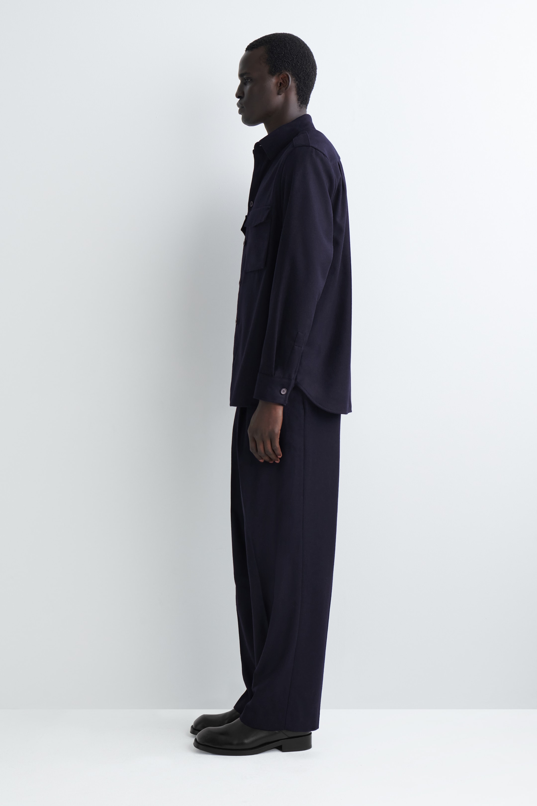 Ingrandisci l'immagine: Un completo composto da una camicia blu scuro e pantaloni a gamba larga viene mostrato in una vista di profilo sinistro. La camicia a maniche lunghe presenta due tasche sul petto con patta e polsini con bottoni, mentre i pantaloni a gamba larga cadono con una vestibilità rilassata e si appoggiano leggermente sulle scarpe eleganti nere.