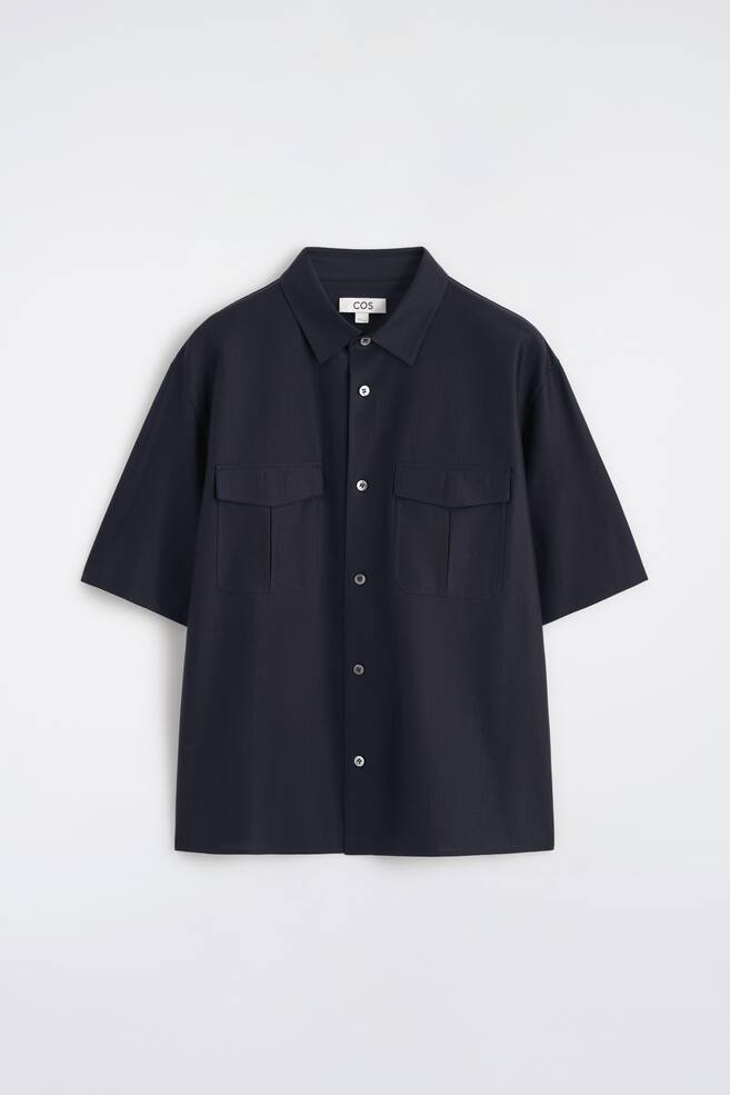 Uma camisa azul-marinho está voltada para a frente, apresentando uma textura subtil de riscas, mangas curtas e uma gola aberta. A camisa exibe uma carcela completa com botões de cor clara e dois bolsos de aba no peito. - 1