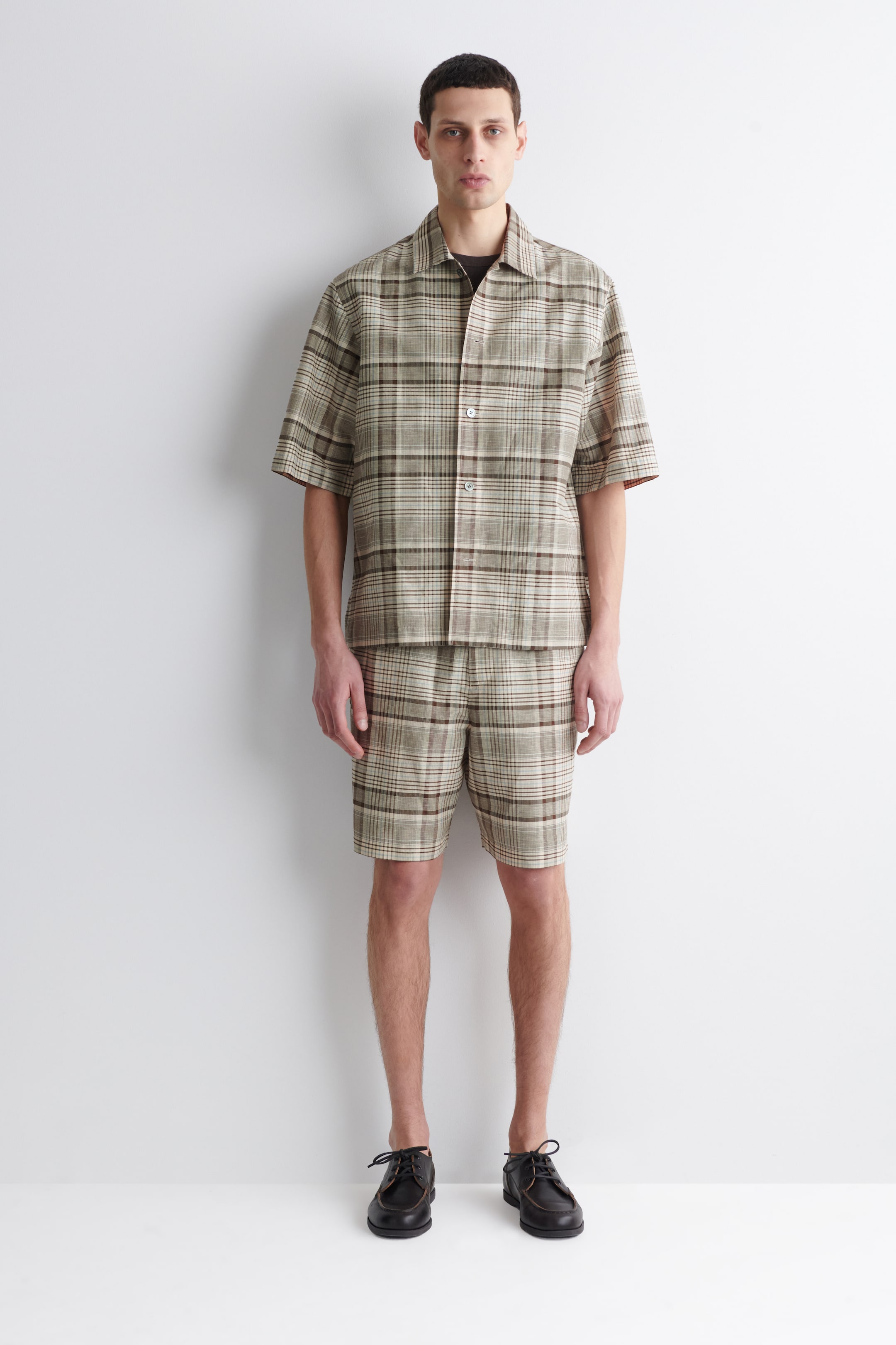 Ver imagem maior: Um conjunto de camisa de manga curta e calções a condizer, com padrão xadrez em tons de bege, castanho e creme, está a ser usado, virado para a frente. A camisa apresenta uma gola aberta e fecho de botões, e está sobreposta a uma camisola interior escura, enquanto os calções têm um comprimento acima do joelho.