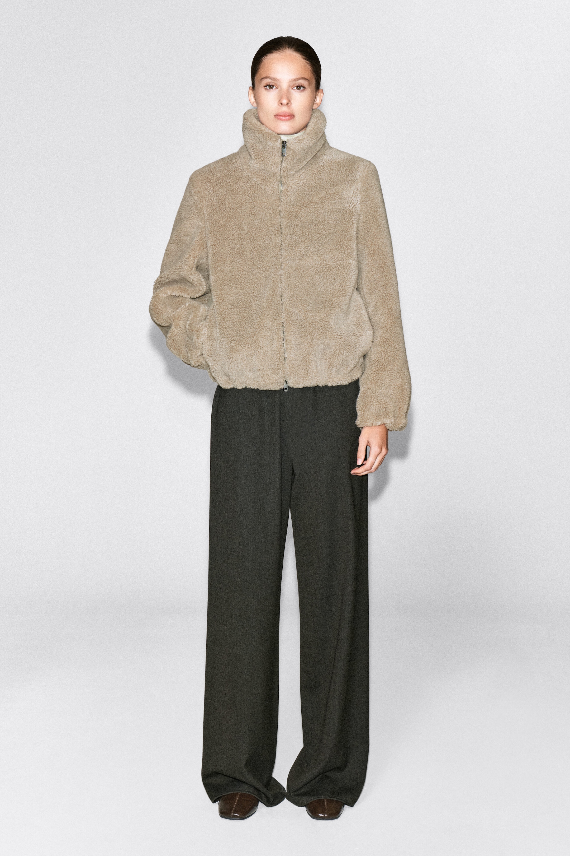 A beige funnel-neck teddy jacket with a textured fleece finish and a front zipper is worn by the model. The jacket is styled with dark wide-leg trousers, and the model rests one hand in the side pocket.