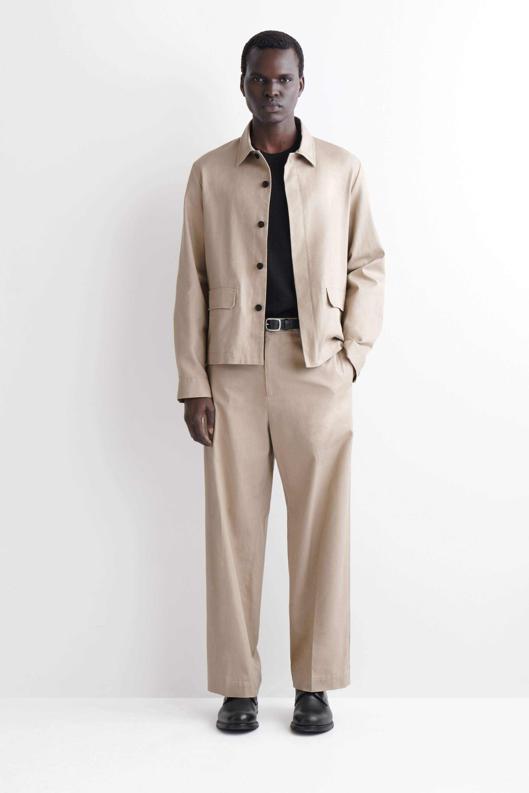 הצגת תמונה גדולה יותר: חליפת שני חלקים בצבע בז' מוצגת על אדם העומד מול המצלמה, וכוללת ז'קט עם צווארון, כפתורים מקדימה ושני כיסים חיצוניים, ומכנסיים רחבים. הז'קט מונח מעל חולצה שחורה, בעוד חגורה שחורה מהדקת את המכנסיים, המשתלבים עם נעליים שחורות עם שרוכים.
