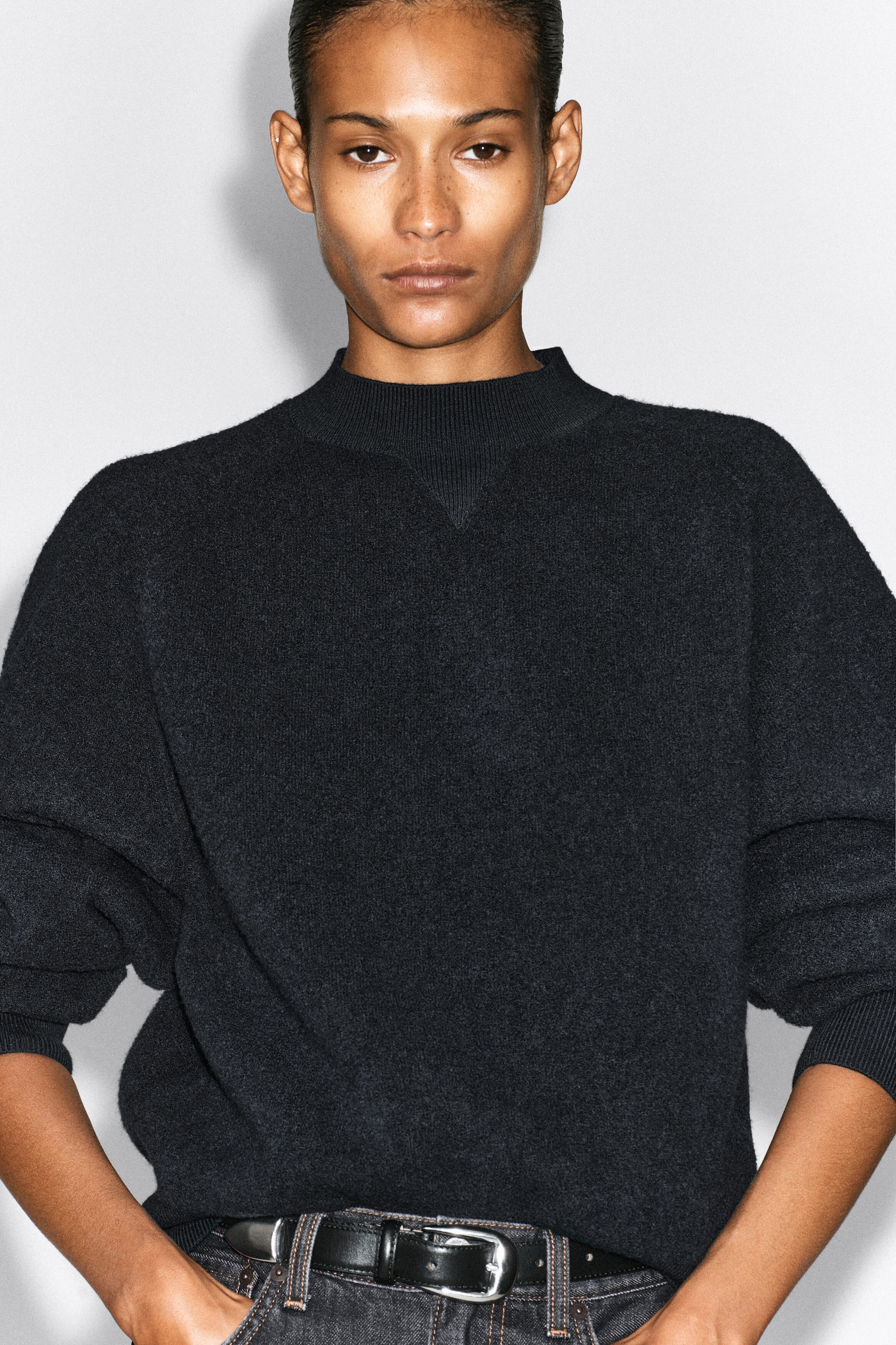 A dark gray, textured knit sweater featuring a ribbed mock neck with a triangular detail is being worn by the model. The relaxed-fit sweater is styled tucked into dark denim jeans secured with a black leather belt. A dark gray, textured knit sweater featuring a ribbed mock neck with a triangular detail is being worn by the model. The relaxed-fit sweater is styled tucked into dark denim jeans secured with a black leather belt.