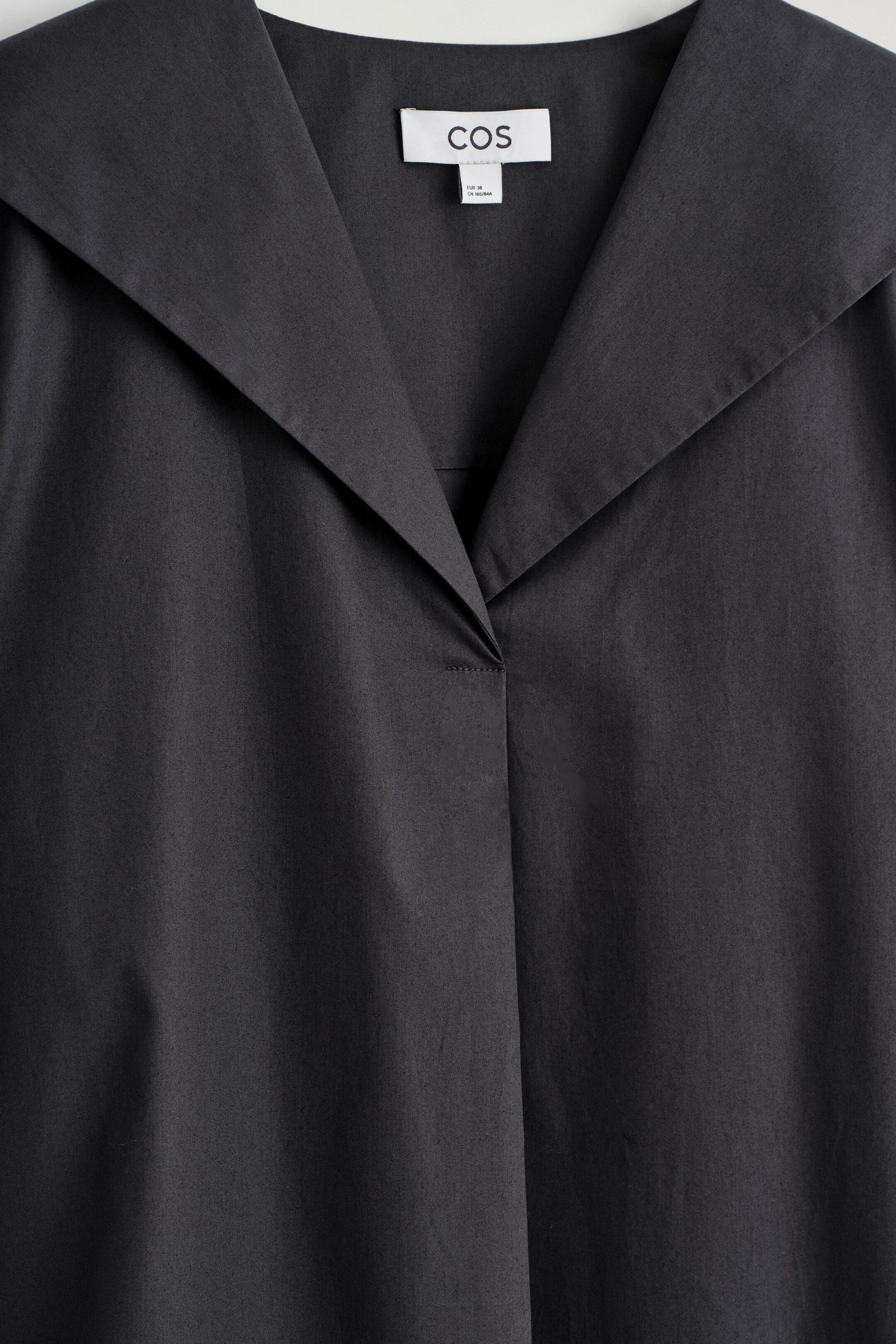 Grösseres Bild ansehen: Ein gewaschenes schwarzes Baumwoll-Poplin-Kleid wird von vorne gezeigt, mit einem breiten Matrosenkragen, der sich über die Schultern erstreckt. Sein V-Ausschnitt bildet einen subtilen Punkt mit einem kleinen Stichdetail.