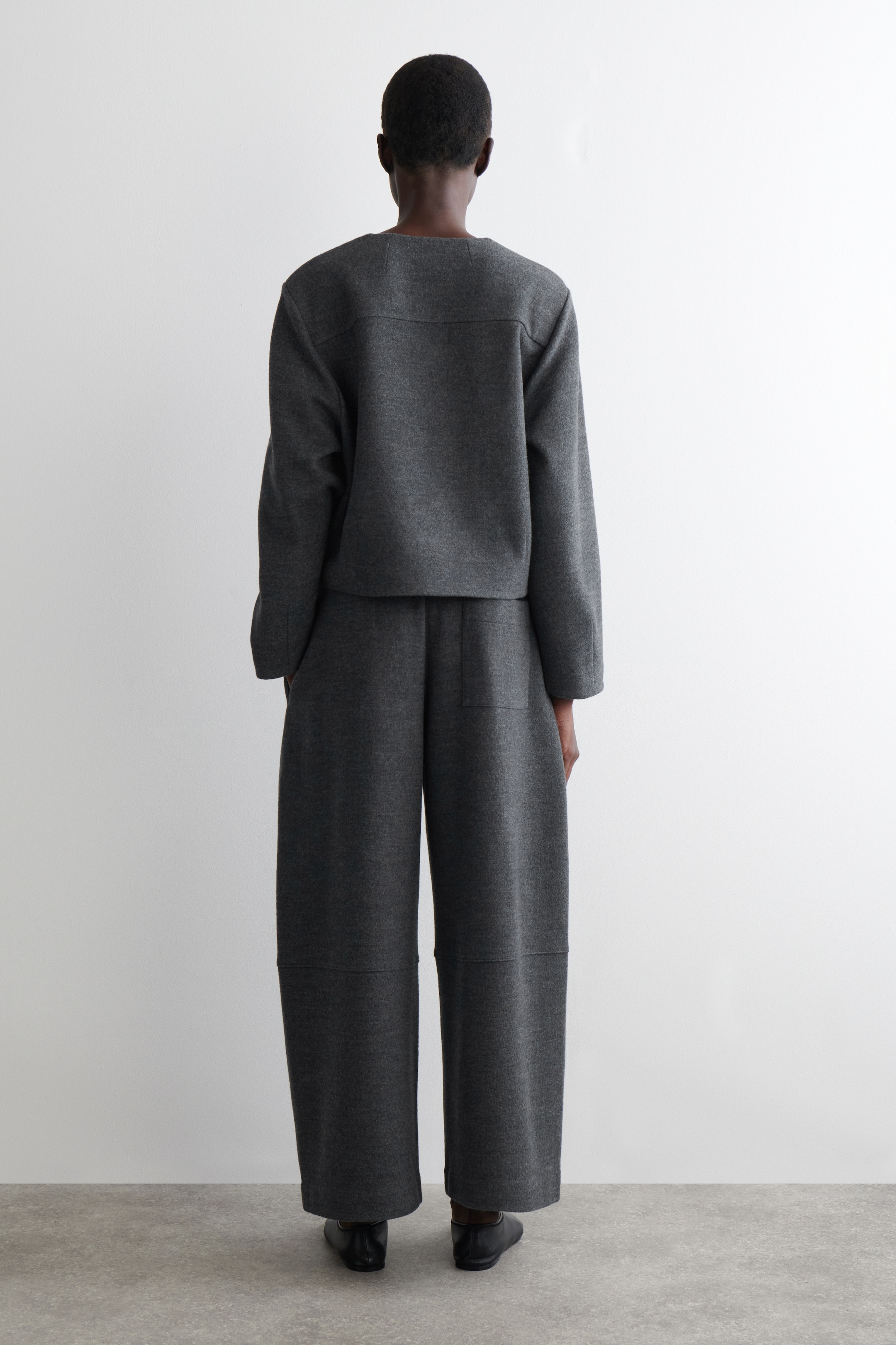 View larger image: A dark grey, structured two-piece outfit, featuring a boxy, long-sleeved top and wide-leg pants, is being displayed from the back. The top is designed with a horizontal seam across the upper back, and the pants are showcasing horizontal seam details on the lower legs and visible side pockets.