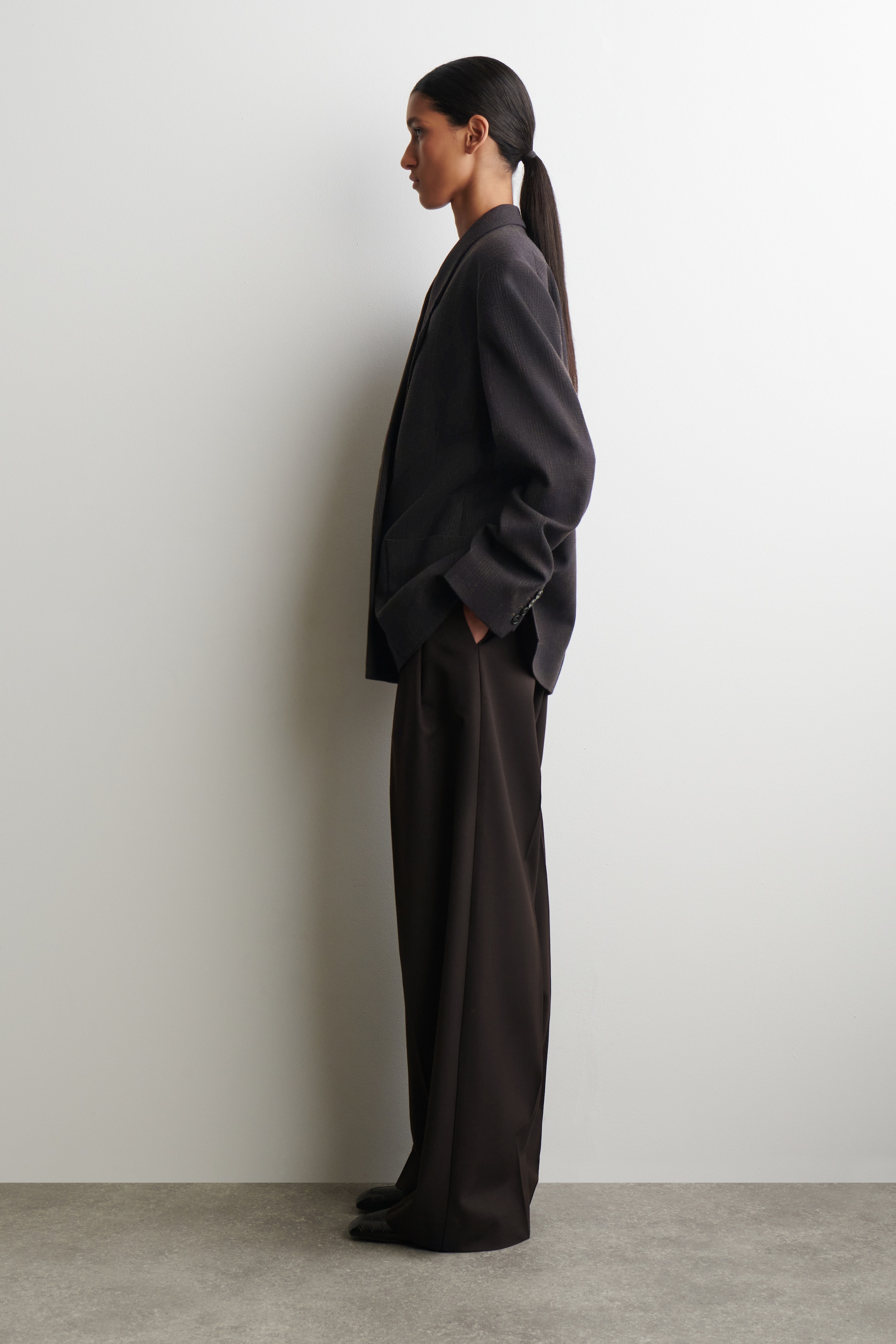 Se større billede: En mørk, subtilt stribet oversized blazer bæres sammen med matchende bukser med brede ben, præsenteret fra en sideprofil. Blazeren har notch-revers og knappede manchetter, mens bukserne har læg.