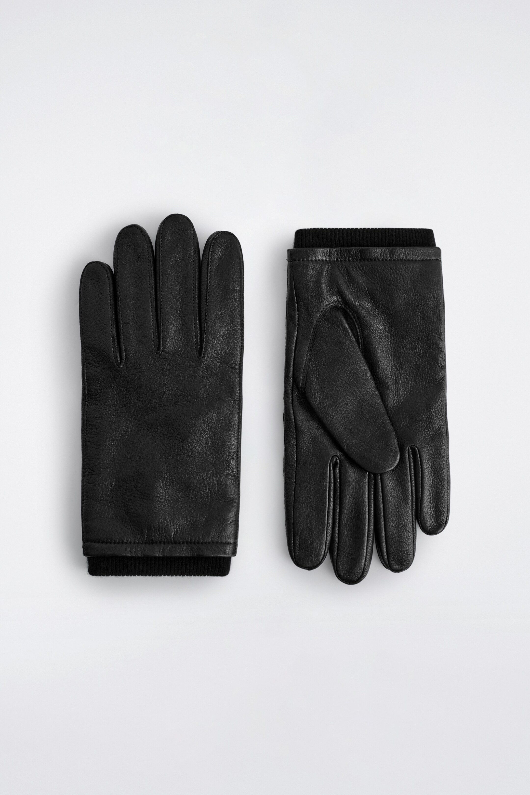 View larger image: A pair of black leather gloves is lying flat, one displaying its palm side and the other its back-of-hand side. Both gloves are featuring black ribbed cuffs.