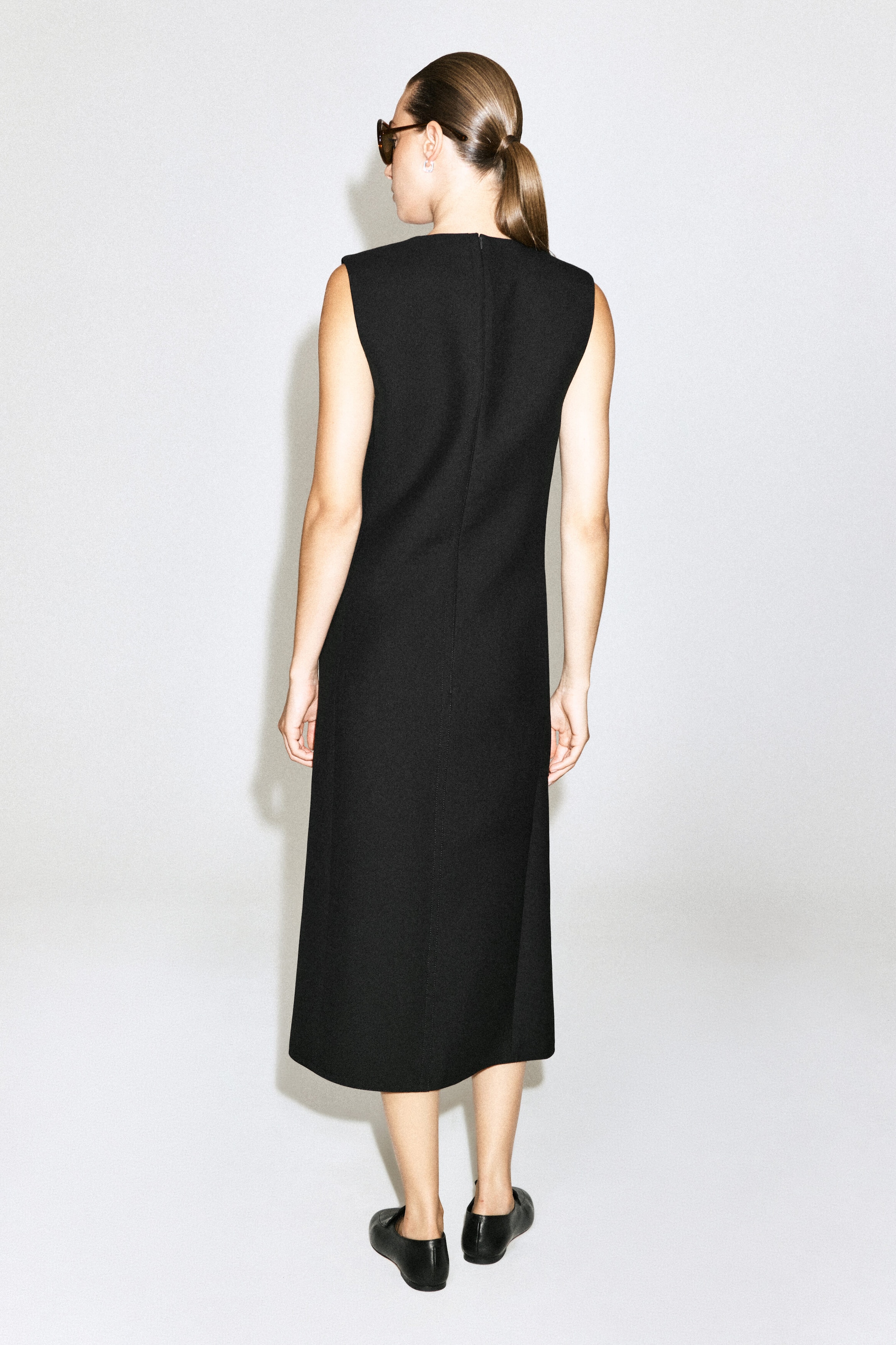 The model is wearing a black sleeveless wool midi dress, highlighting its straight silhouette, high back neckline, and center back seam. The dress is styled with flat black leather shoes.