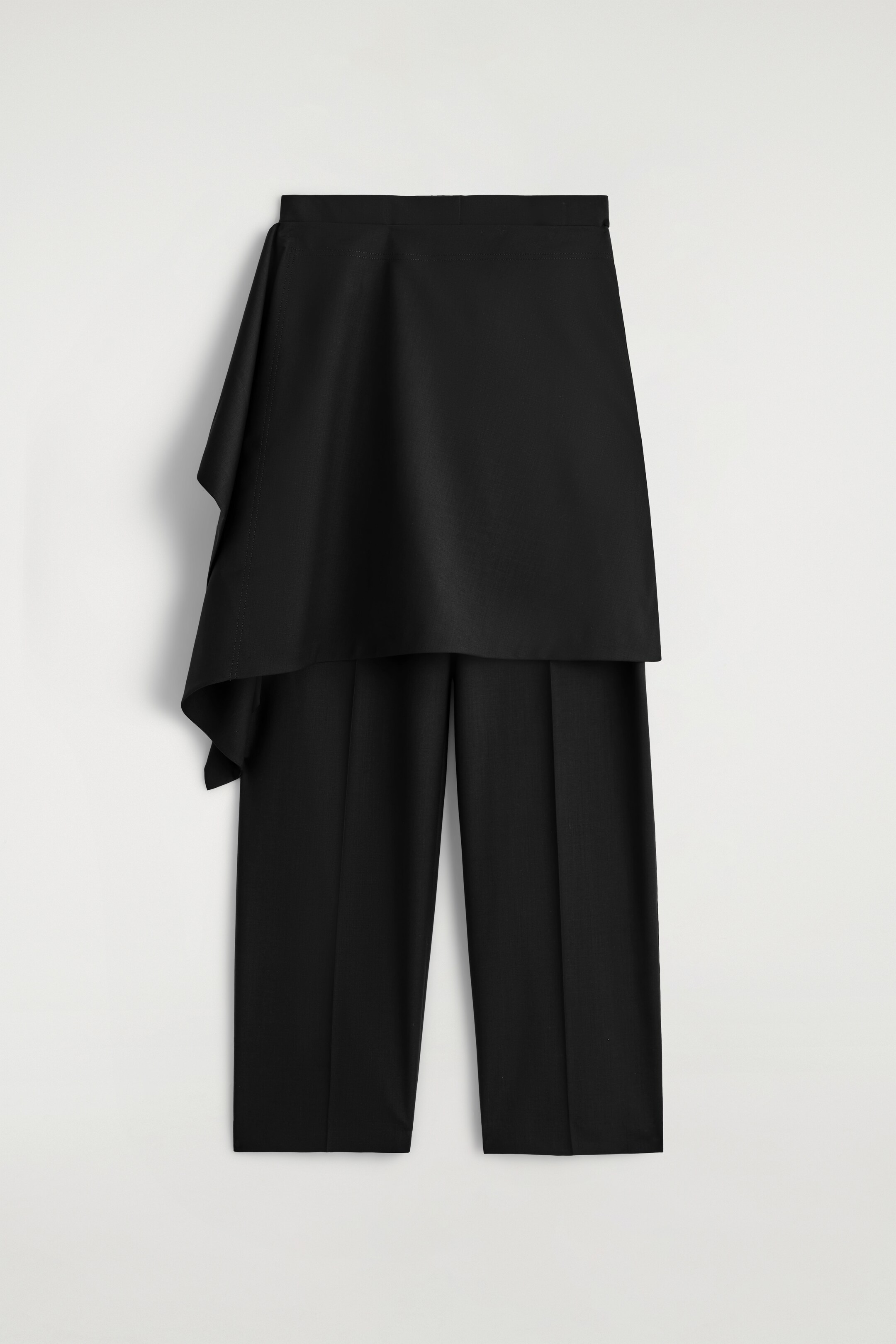 Agrandir l'image: Un pantalon noir est présenté de FACE, avec une surjupe drapée qui s'enroule autour de la taille et tombe fluidement sur le côté gauche. Le pantalon présente des plis marqués à l'avant.