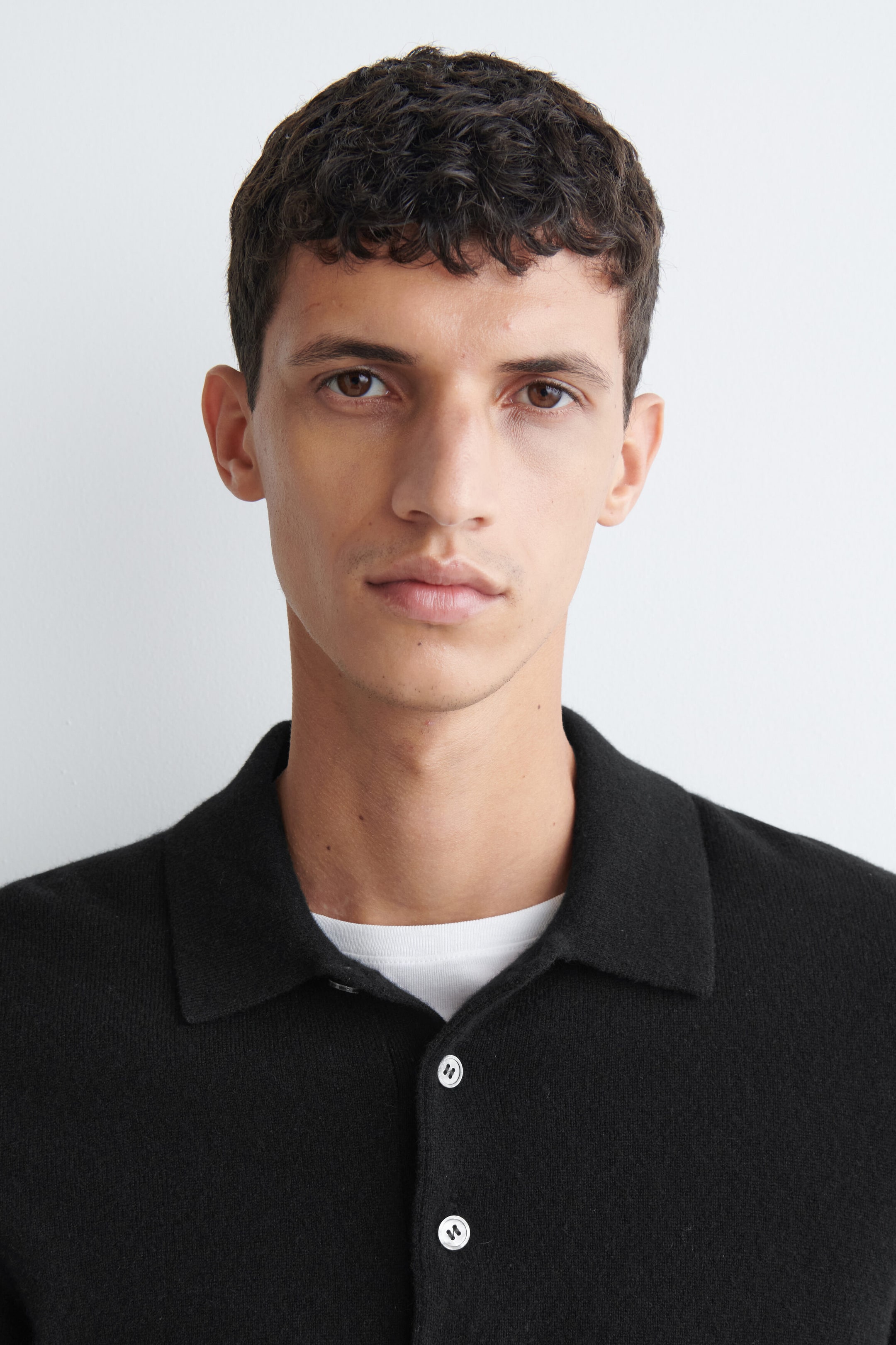 Una polo in maglia nera con colletto classico e bottoni bianchi è indossata dalla modella. La polo è sovrapposta a una T-shirt bianca girocollo, visibile allo scollo. Una polo in maglia nera con colletto classico e bottoni bianchi è indossata dalla modella. La polo è sovrapposta a una T-shirt bianca girocollo, visibile allo scollo.
