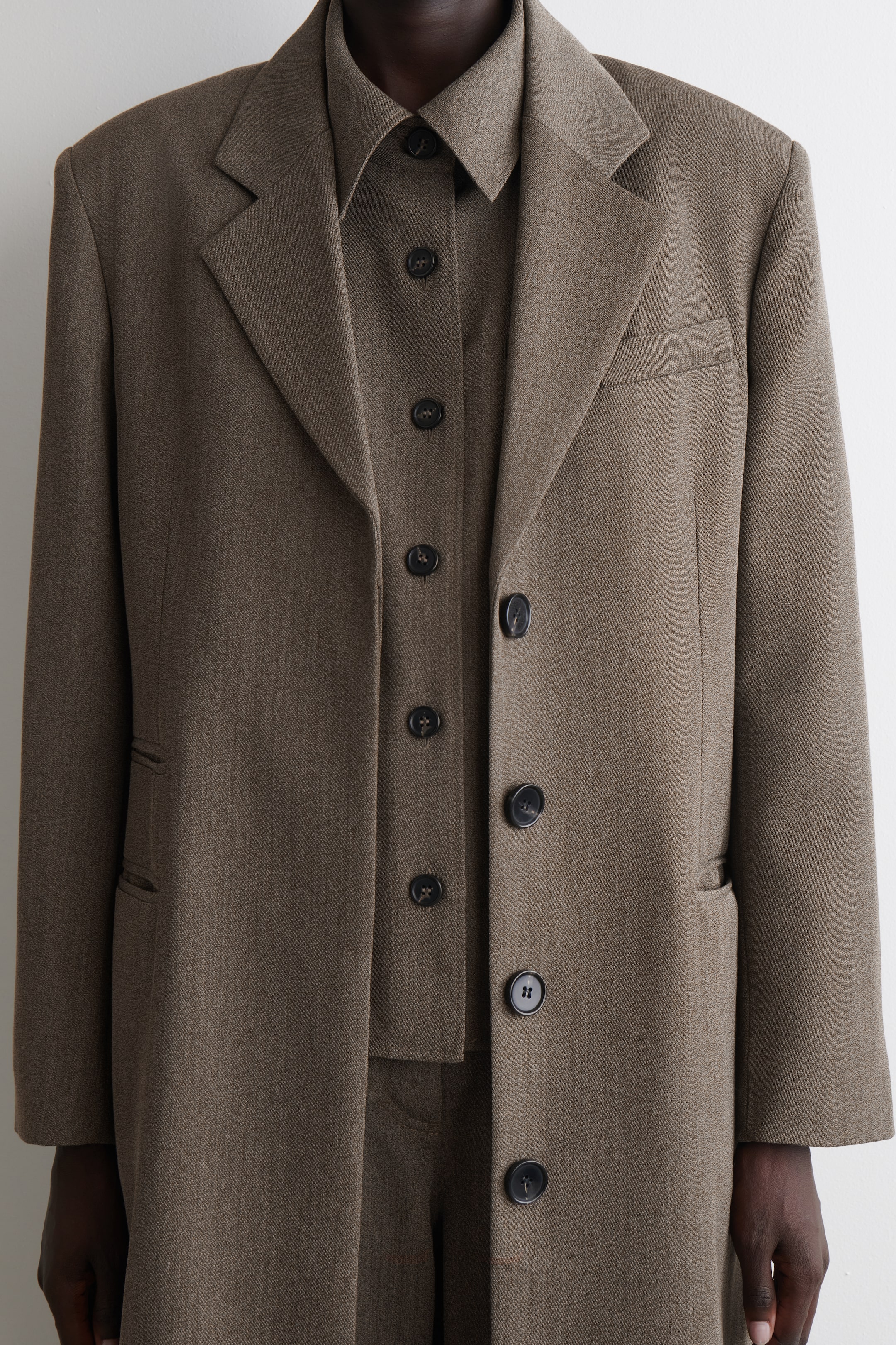 Ingrandisci l'immagine: Un abito marrone è presentato frontalmente, caratterizzato da un blazer oversize con ampi revers a tacca, una tasca sul petto e due tasche con patta. Il blazer, chiuso da uno dei suoi quattro bottoni frontali scuri, è stratificato su una camicia con colletto coordinata, con entrambi i capi realizzati in un tessuto marrone testurizzato.