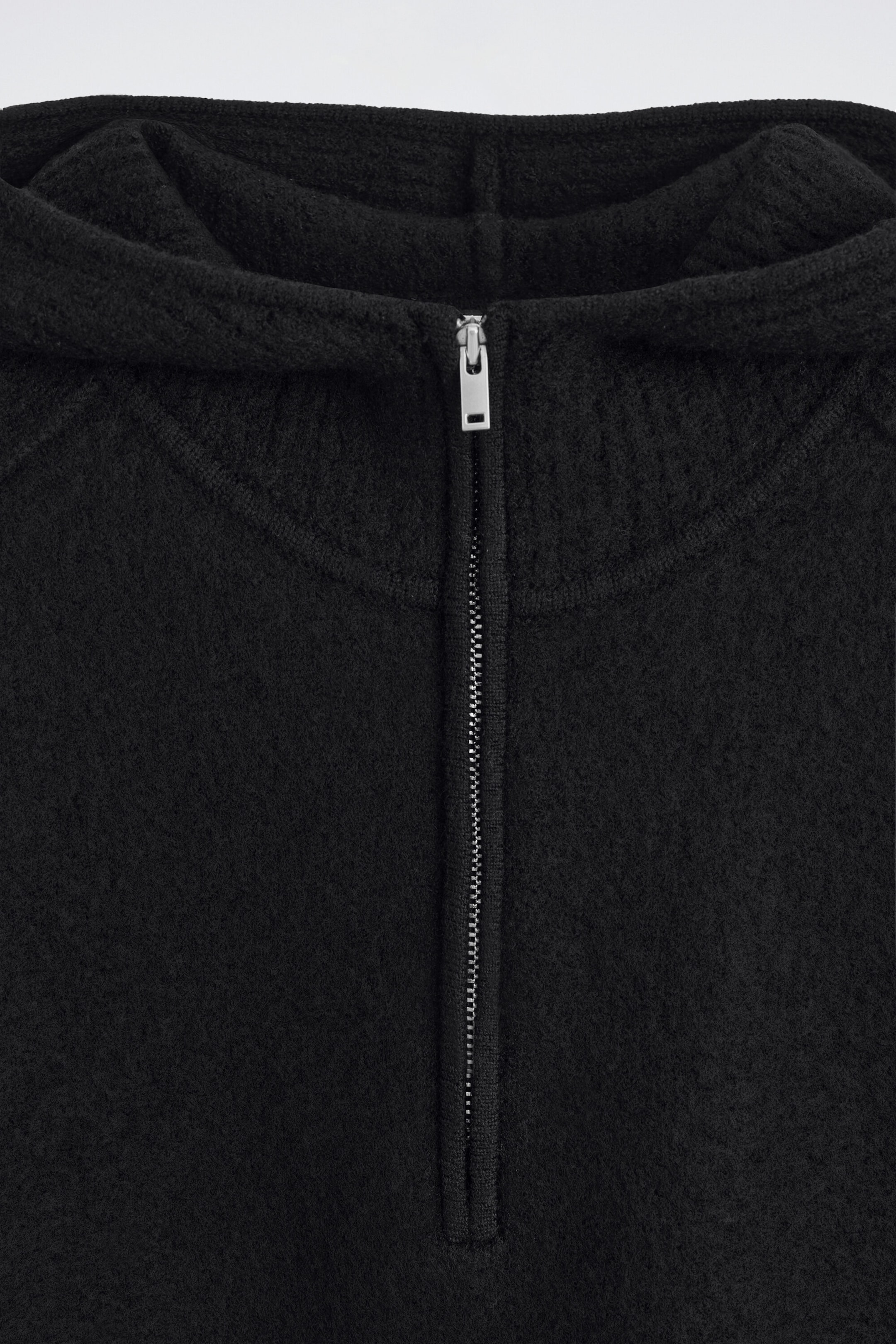 A close-up front view showcases a black merino wool half-zip hoodie with a textured knit finish. The garment features a silver metal half-zip fastening with a rectangular pull tab, alongside a ribbed collar detail and an attached hood. A close-up front view showcases a black merino wool half-zip hoodie with a textured knit finish. The garment features a silver metal half-zip fastening with a rectangular pull tab, alongside a ribbed collar detail and an attached hood.