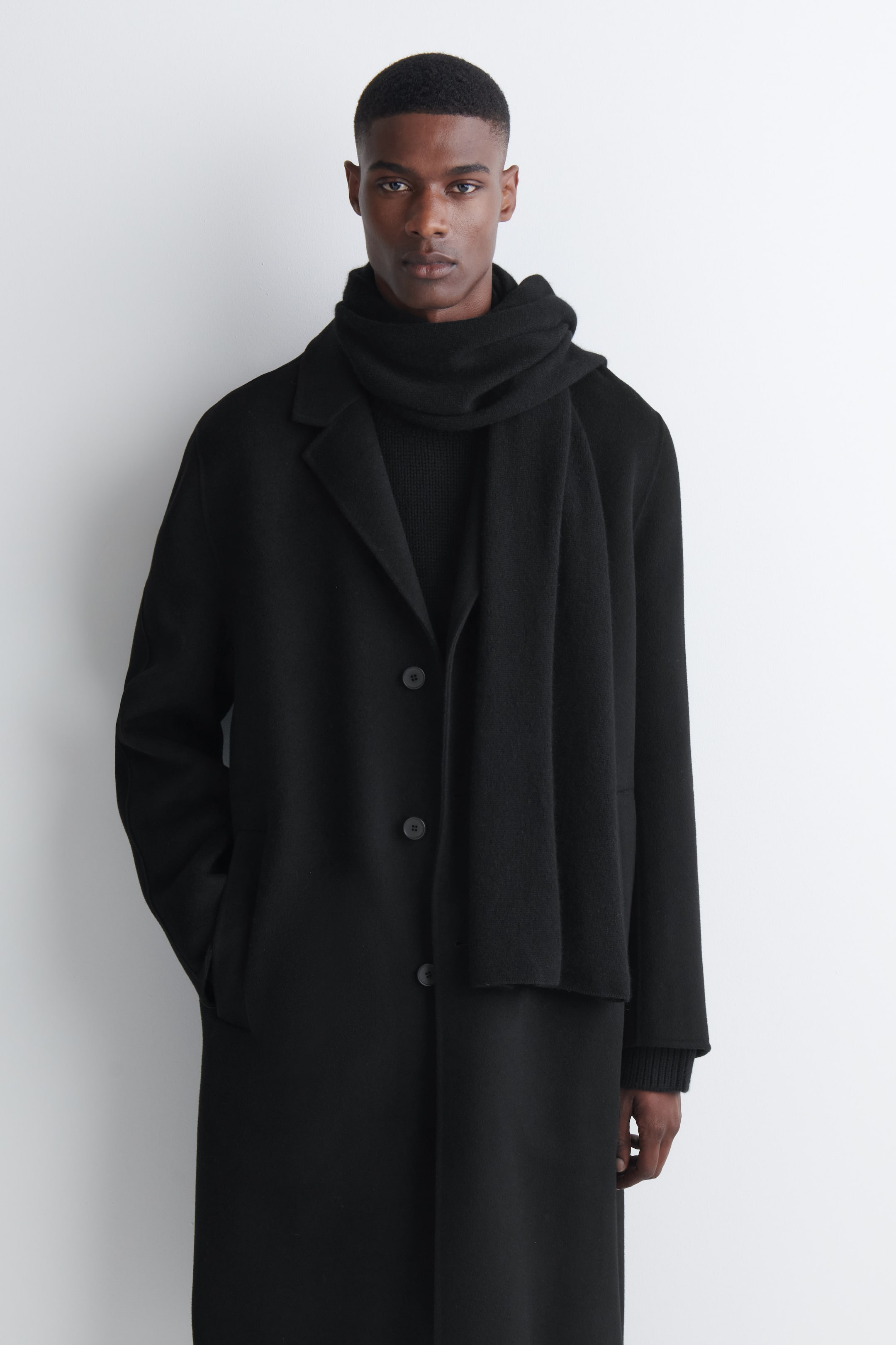 View larger image: The model is wearing a black cashmere scarf wrapped around the neck, with one end draped down the front. The scarf is layered over a matching black single-breasted overcoat, highlighting its soft texture.