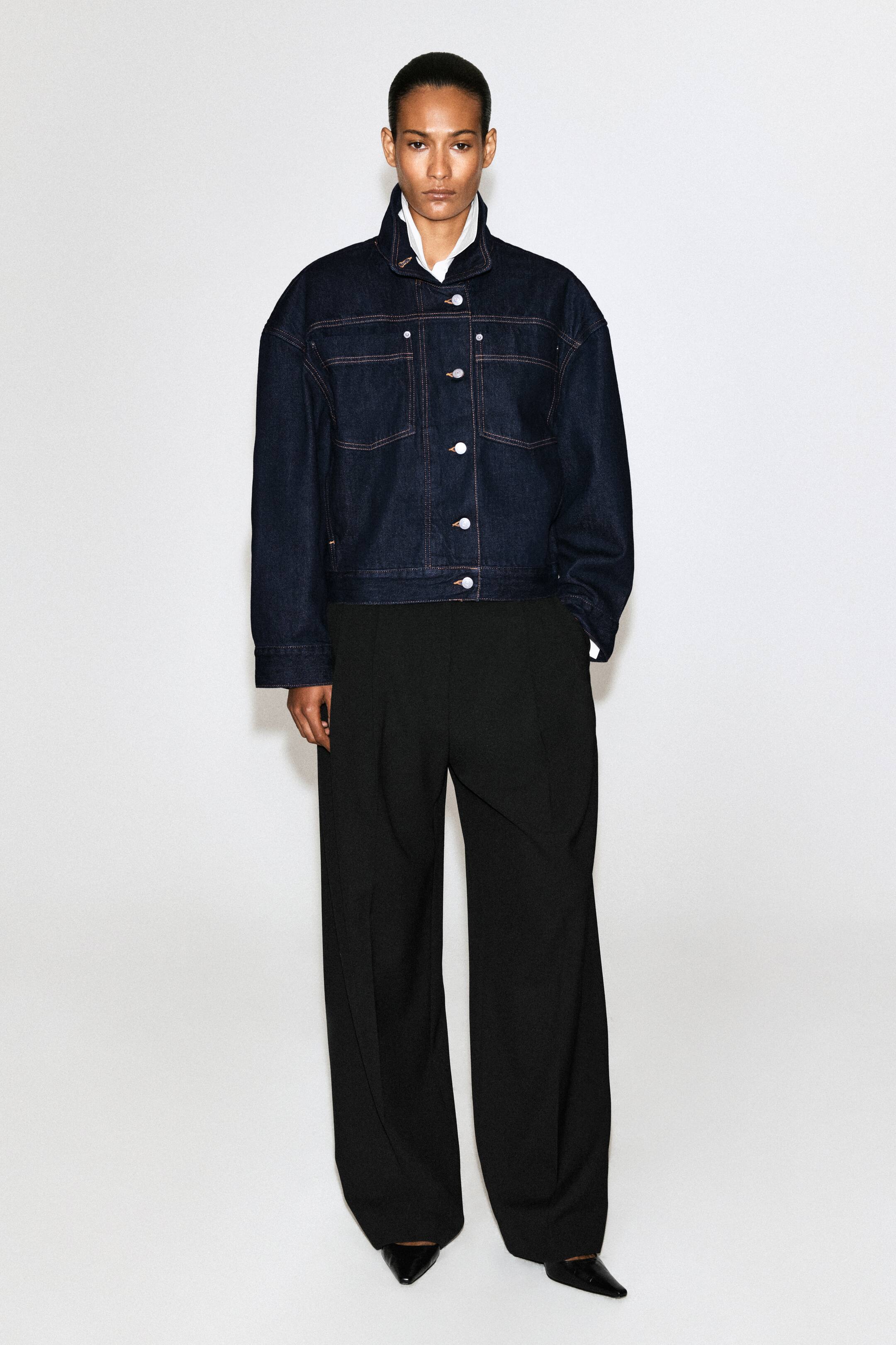 View larger image: A dark blue denim jacket with a relaxed fit is being worn by the model. It features oversized patch pockets and white button fastenings, and its exaggerated pointed collar is styled upwards as a funnel neck over a white shirt.