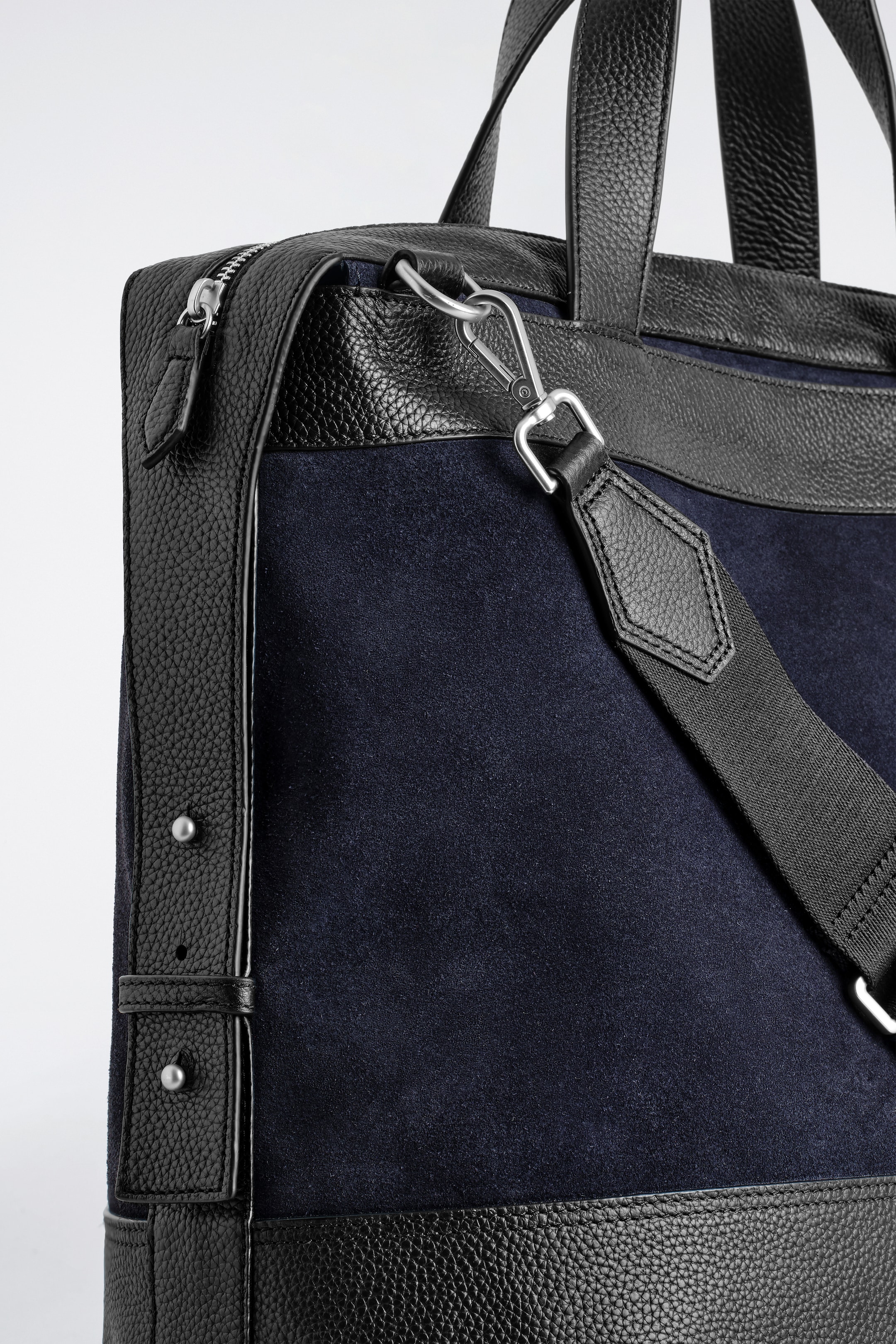 Visualizza immagine più grande: Una borsa in camoscio blu scuro presenta accenti in pelle nera a grana lungo i bordi e i pannelli laterali. Una chiusura con cerniera color argento è visibile in alto a sinistra, e una tracolla in tessuto nero è attaccata con un moschettone d'argento al lato destro della borsa.
