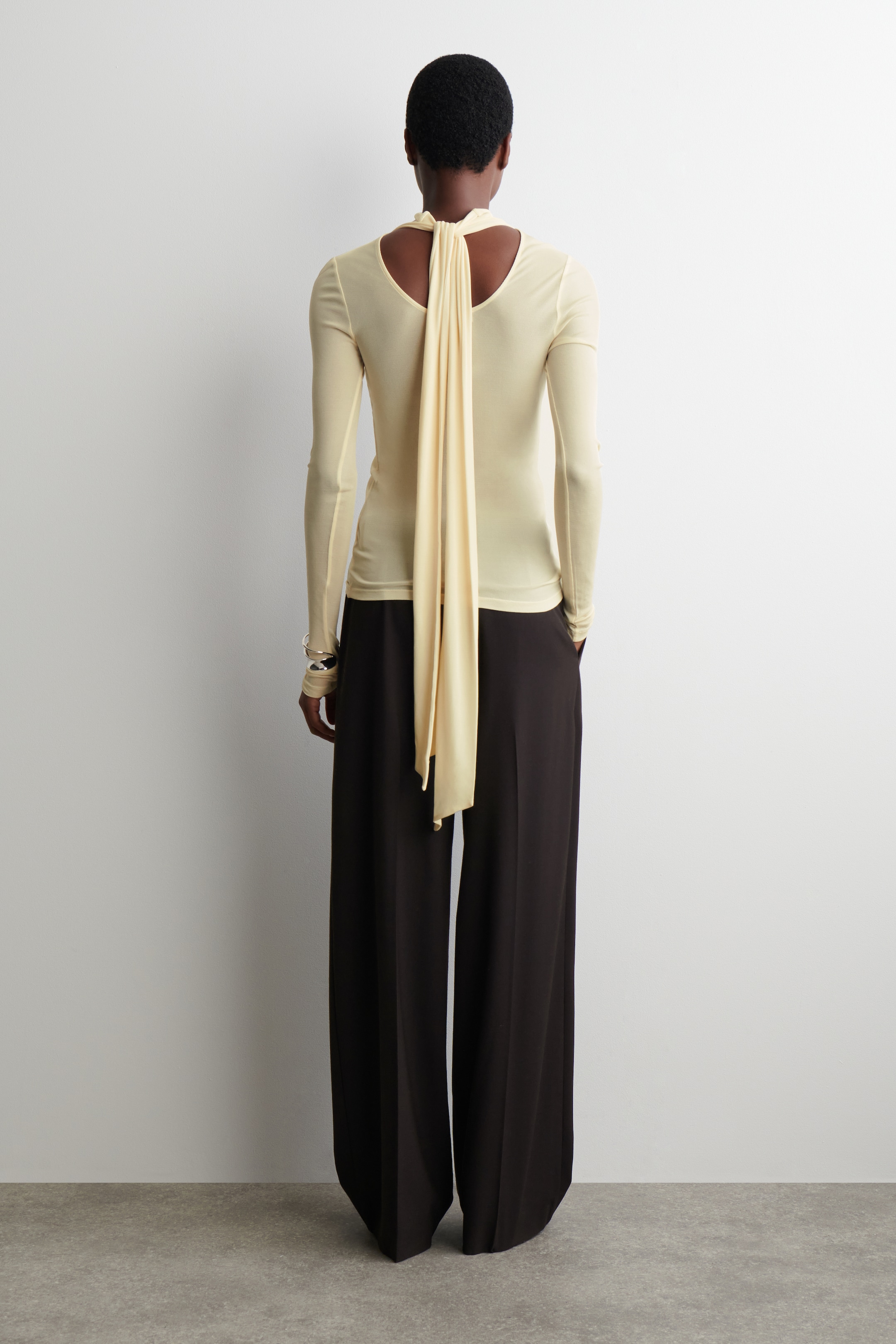Agrandir l'image: Un haut ajusté à manches longues de couleur jaune pâle est présenté de dos, avec un détail découpé au niveau du haut du dos et un long lien en tissu qui descend. Ce haut est assorti à un pantalon large et foncé.