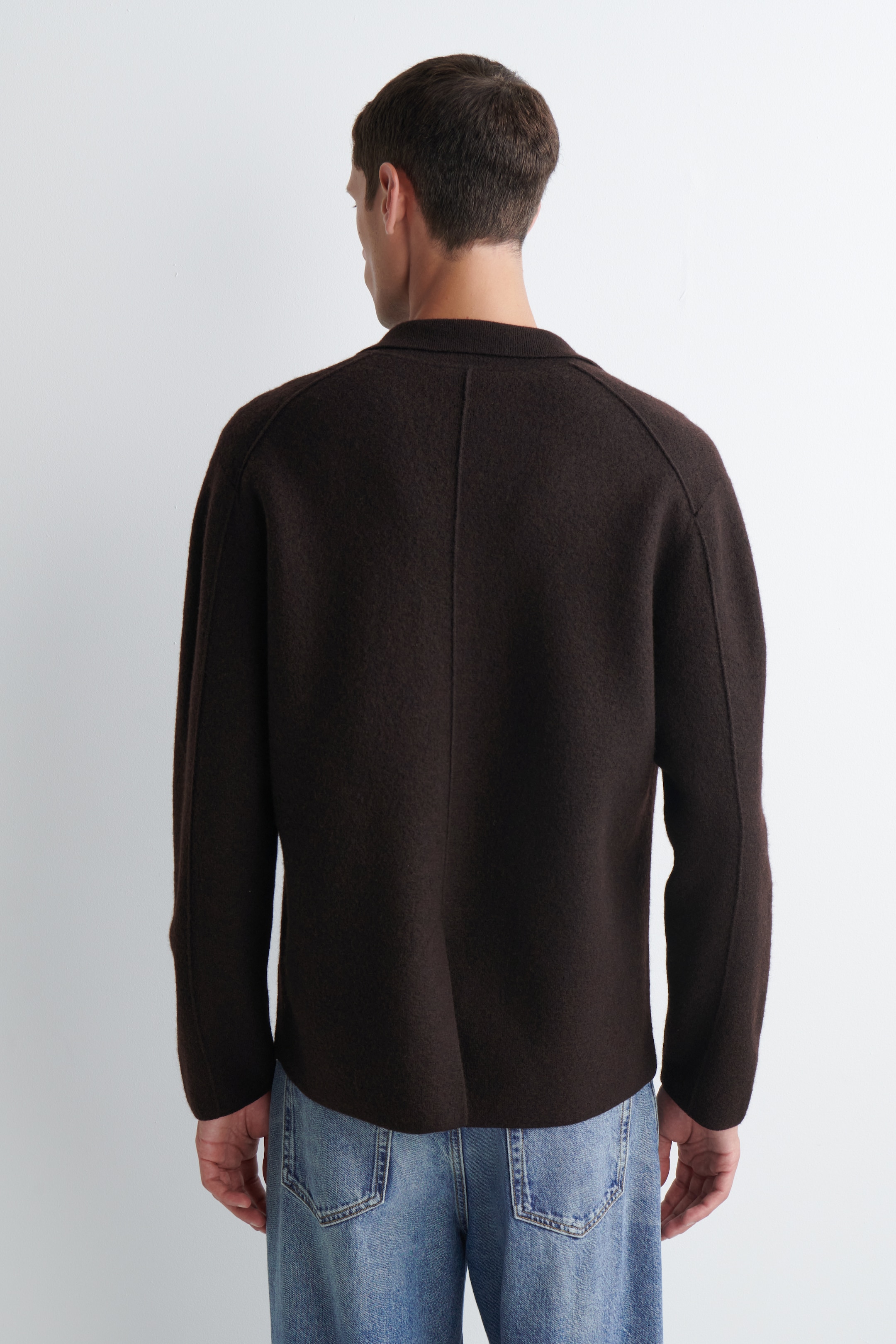Größeres Bild ansehen: Eine dunkelbraune Jacke wird von hinten präsentiert, um ihre lockere Passform und den strukturierten Stoff zu zeigen. Sie verfügt über einen kurzen Stehkragen, eine markante vertikale Naht in der Mitte des Rückens und Raglanärmel mit zusätzlichen vertikalen Nähten. Der Saum der Jacke ruht knapp über einer hell gewaschenen Jeans.