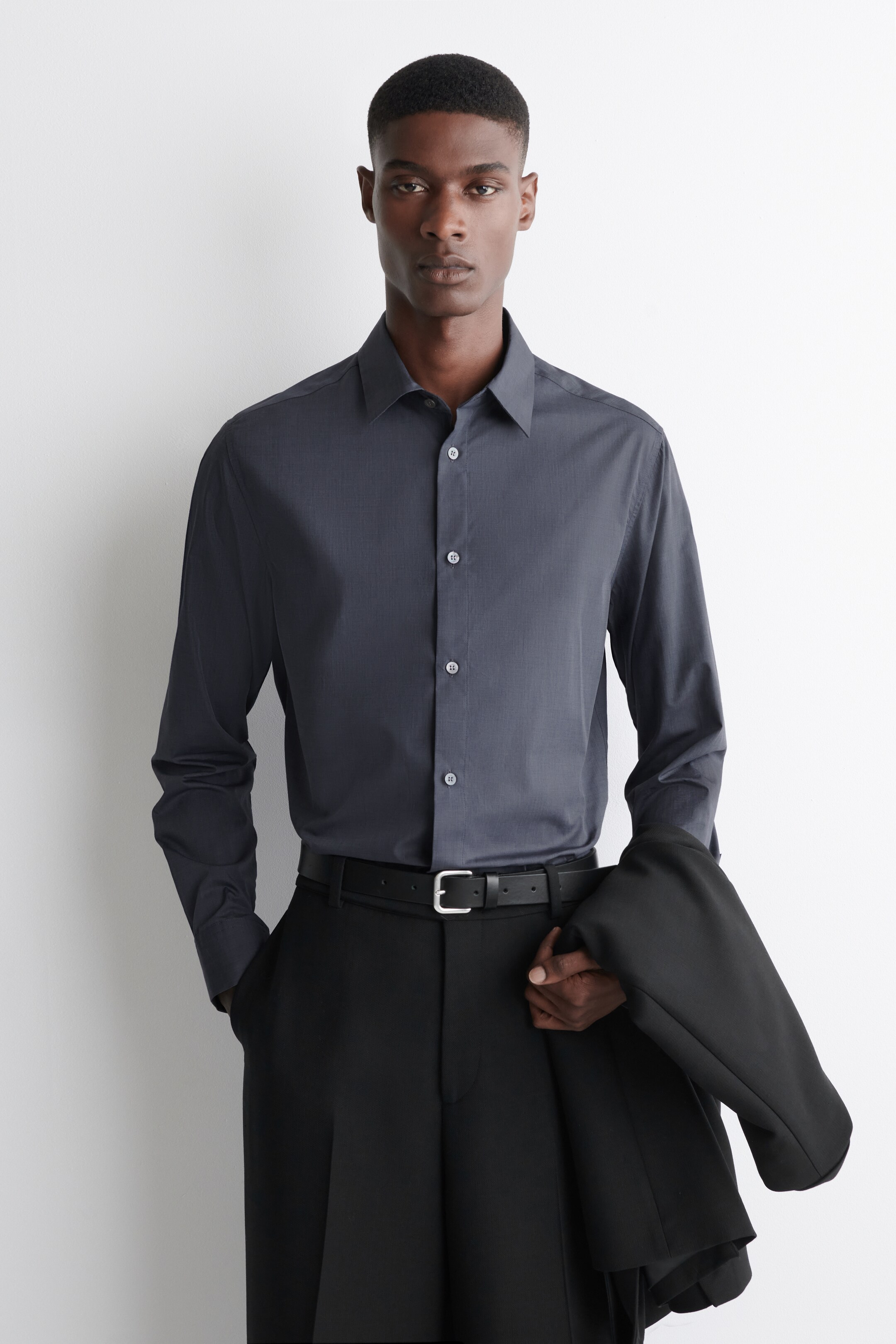 Agrandir l'image: Une chemise gris foncé avec un tissage fil-à-fil et un col pointu est portée par le modèle. Elle est dotée de boutons de couleur claire sur le devant et est assortie à un pantalon noir.