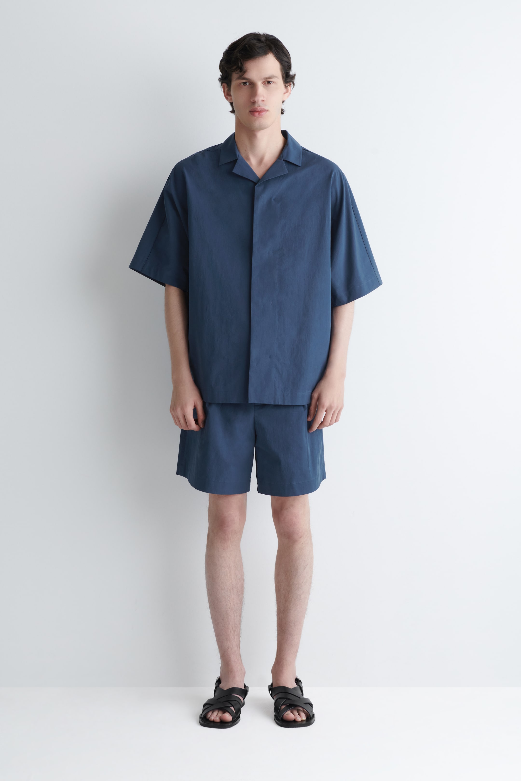 Grotere afbeelding bekijken: Een donkerblauw shirt met korte mouwen wordt gedragen met bijpassende shorts met een ontspannen pasvorm, gepresenteerd aan de voorkant. Het shirt heeft een open kampkraag en brede mouwen, terwijl de shorts tot halverwege de dij komen.