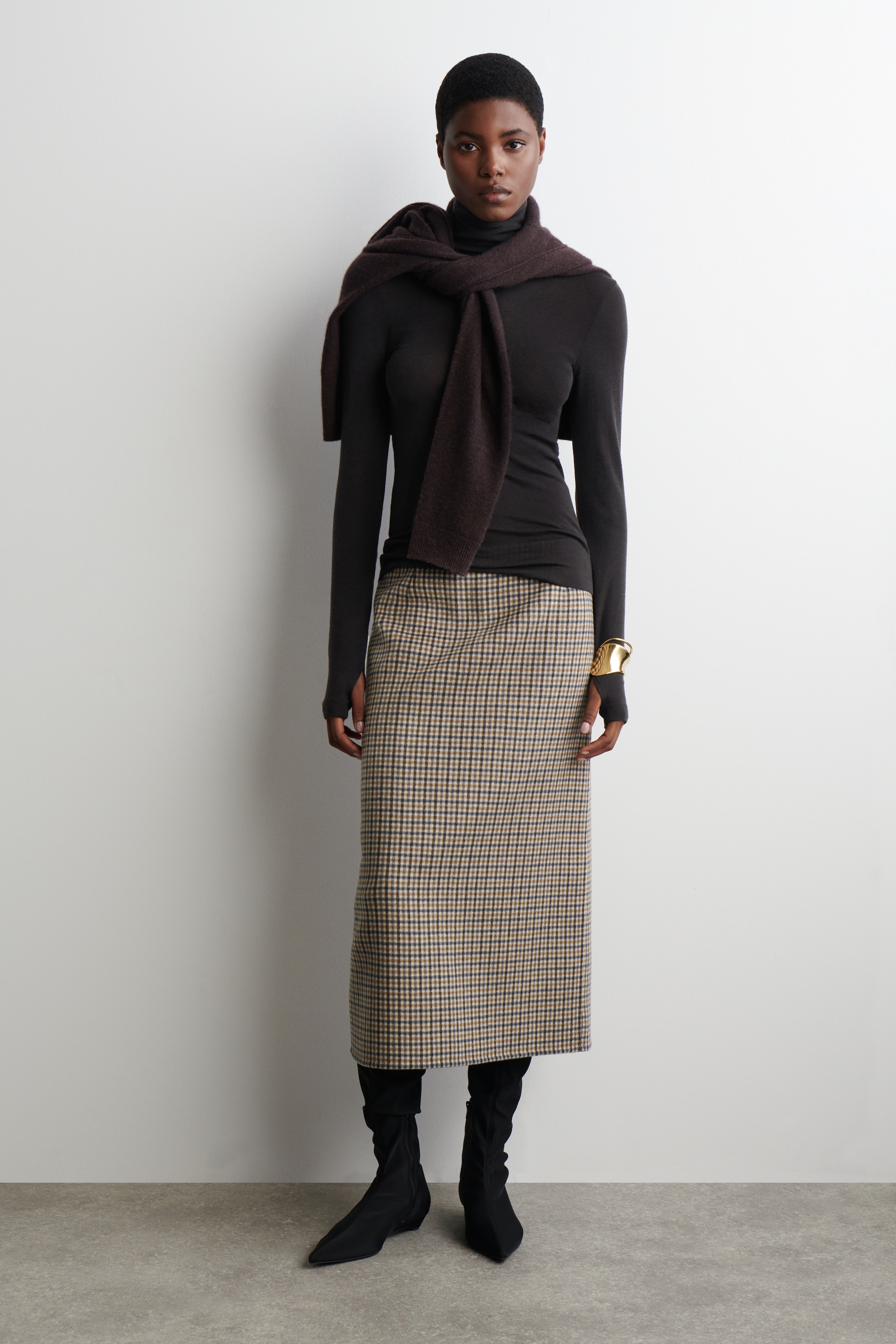 Agrandir l'image: Un haut à col roulé noir, ajusté, avec des poignets à trous pour les pouces est stylisé avec un pull en tricot marron foncé drapé sur les épaules, et une jupe midi à coupe droite avec un motif à carreaux beige et noir. L'ensemble est complété par des bottes noires à bout pointu, mi-mollet, et un bracelet manchette doré est visible sur le poignet droit.