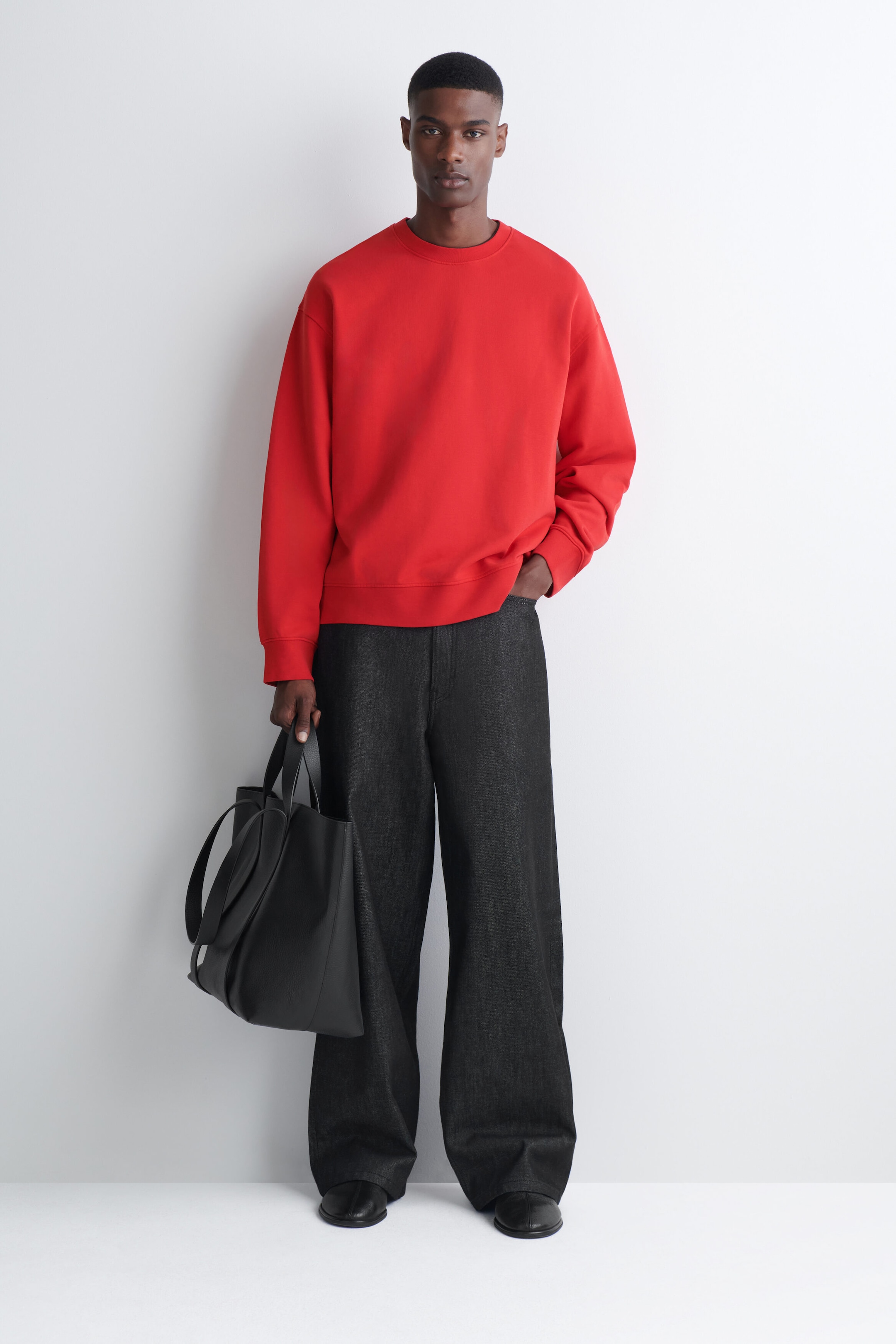 A bright red crew-neck sweatshirt is worn by the model, featuring a relaxed fit with a ribbed collar, cuffs and hem. The sweatshirt is styled with dark grey wide-leg trousers and a black leather tote bag carried in one hand. A bright red crew-neck sweatshirt is worn by the model, featuring a relaxed fit with a ribbed collar, cuffs and hem. The sweatshirt is styled with dark grey wide-leg trousers and a black leather tote bag carried in one hand.