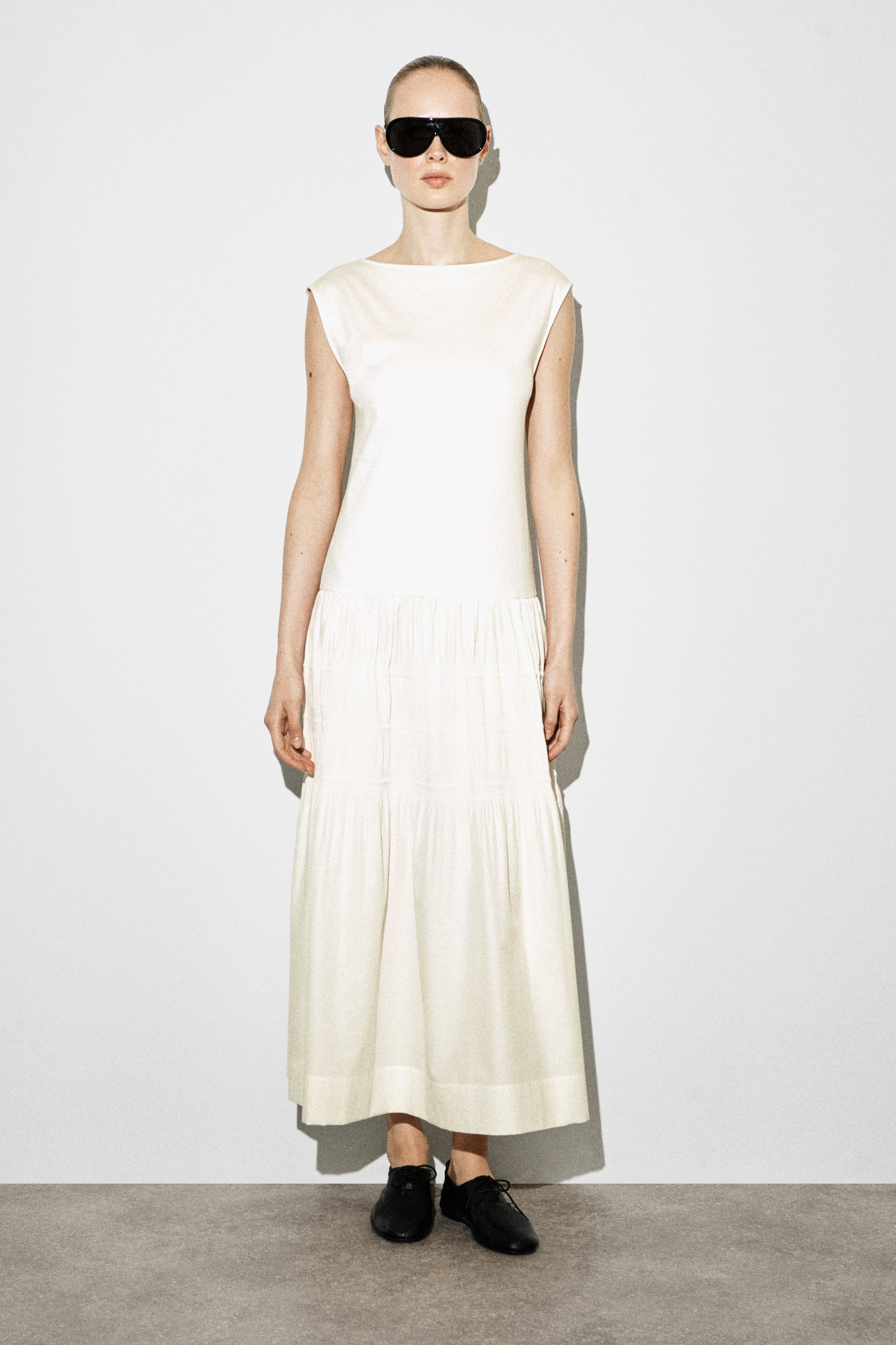 Agrandir l'image: Une robe sans manches de couleur blanc cassé présente un col bateau et un corsage lisse, se transformant en une jupe à volants froncés avec un détail de smocks sur le volant supérieur, s'étendant jusqu'à la cheville.