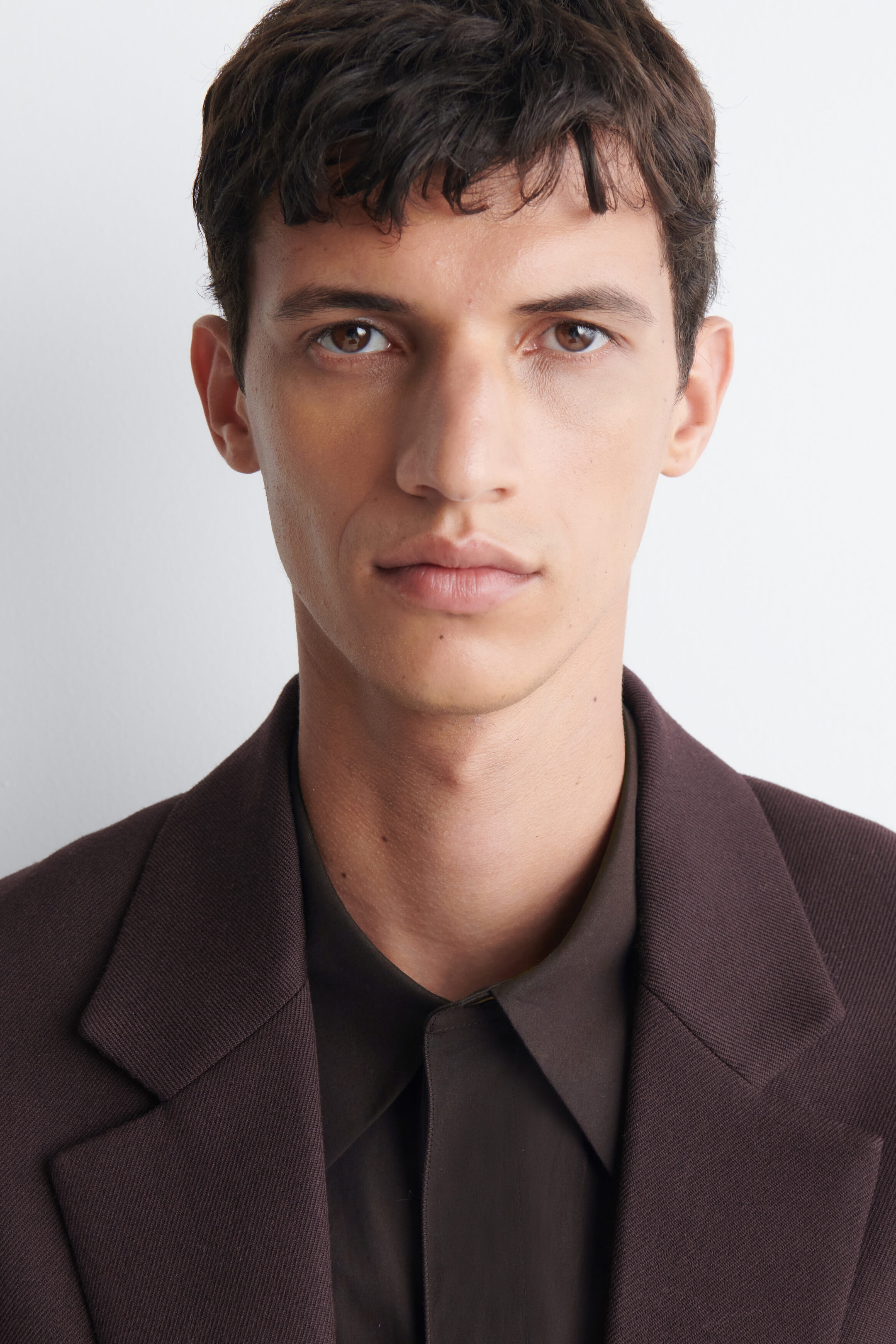 The model is wearing a dark brown blazer, layered over a dark brown collared shirt. The model is wearing a dark brown blazer, layered over a dark brown collared shirt.