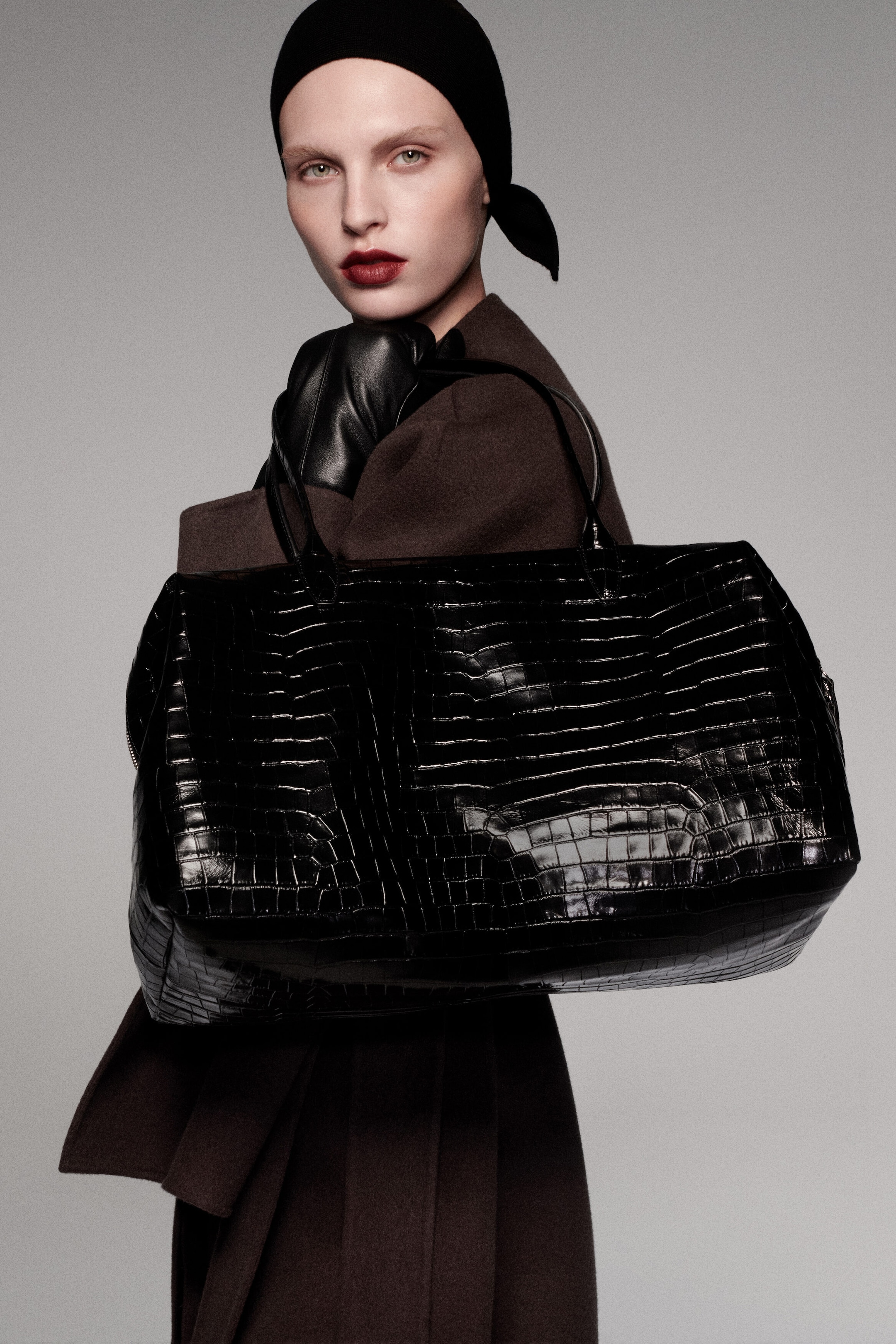 The dark brown double-faced wool-blend cape and belted long coat are shown from a front-facing angle, highlighting their structured draped layers and belted waist. A large, glossy black croc-effect leather Frieze bowling bag is carried over the shoulder by its top handles, showcasing its textured crocodile pattern and spacious design. The dark brown double-faced wool-blend cape and belted long coat are shown from a front-facing angle, highlighting their structured draped layers and belted waist. A large, glossy black croc-effect leather Frieze bowling bag is carried over the shoulder by its top handles, showcasing its textured crocodile pattern and spacious design.