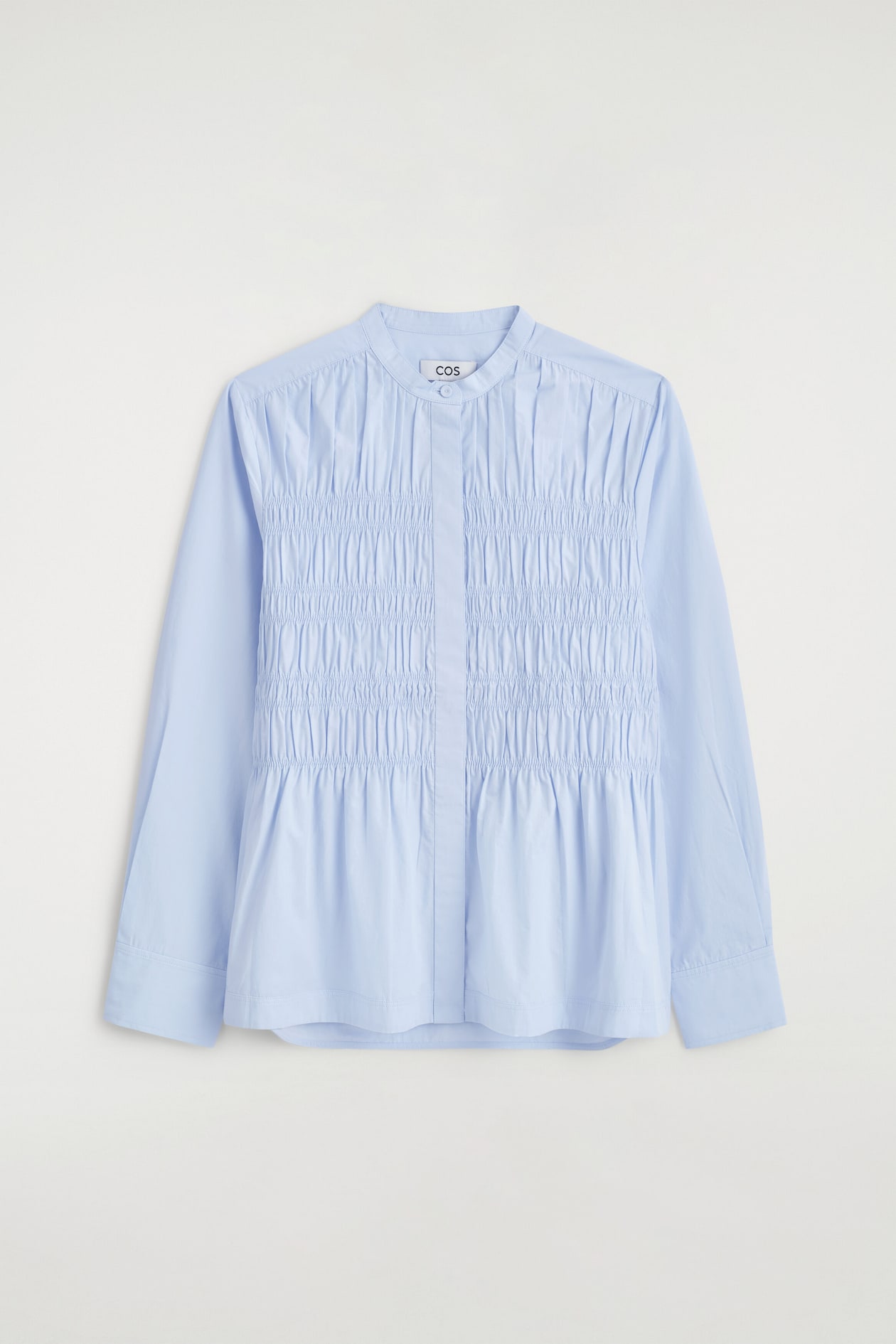 A light blue cotton blouse is shown from the front, featuring a band collar, long sleeves with wide cuffs, and a concealed button placket. The front panel showcases extensive vertical ruching and smocked detailing that falls into a gathered hem.