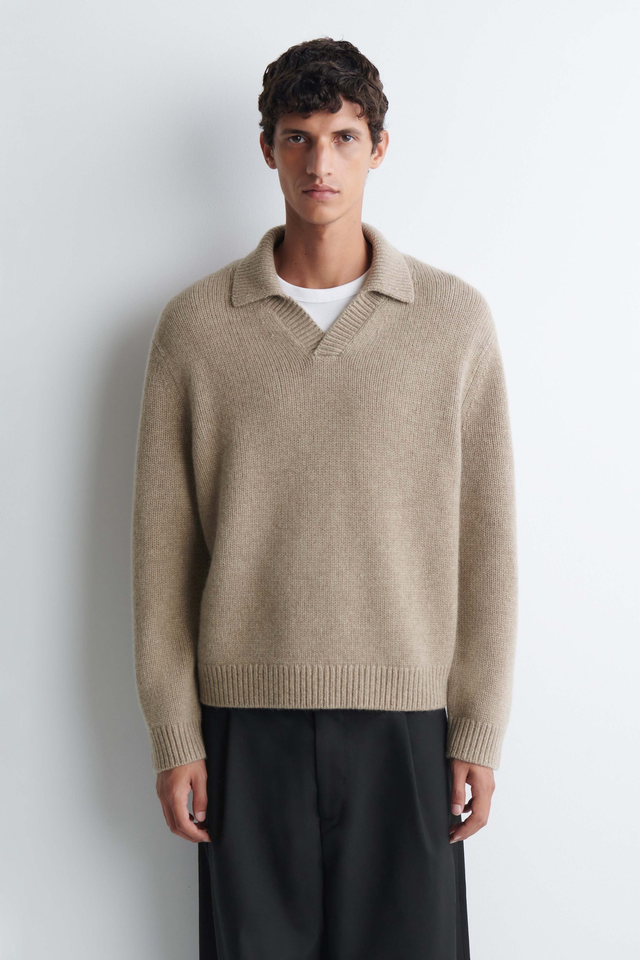 The model is wearing a chunky cashmere polo sweater in light beige, featuring a ribbed knit texture, a spread collar, and an open V-neck. The sweater is layered over a white crewneck T-shirt and styled with black trousers. The model is wearing a chunky cashmere polo sweater in light beige, featuring a ribbed knit texture, a spread collar, and an open V-neck. The sweater is layered over a white crewneck T-shirt and styled with black trousers.