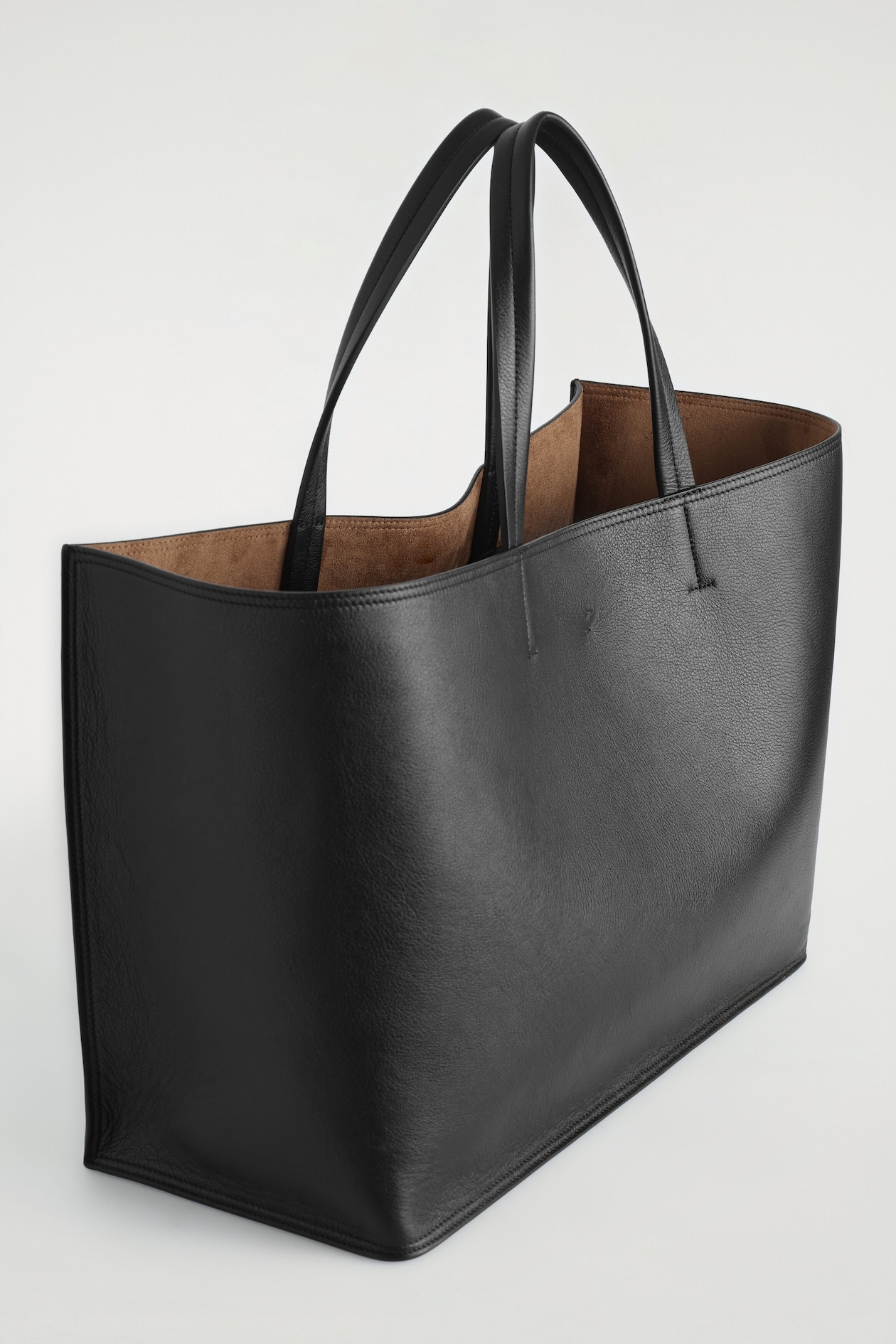 A structured black grained-leather tote bag is standing at an angle, facing right, showcasing its dual top handles and open-top design. The bag displays a contrasting brown suede lining inside its spacious, rectangular compartment.
