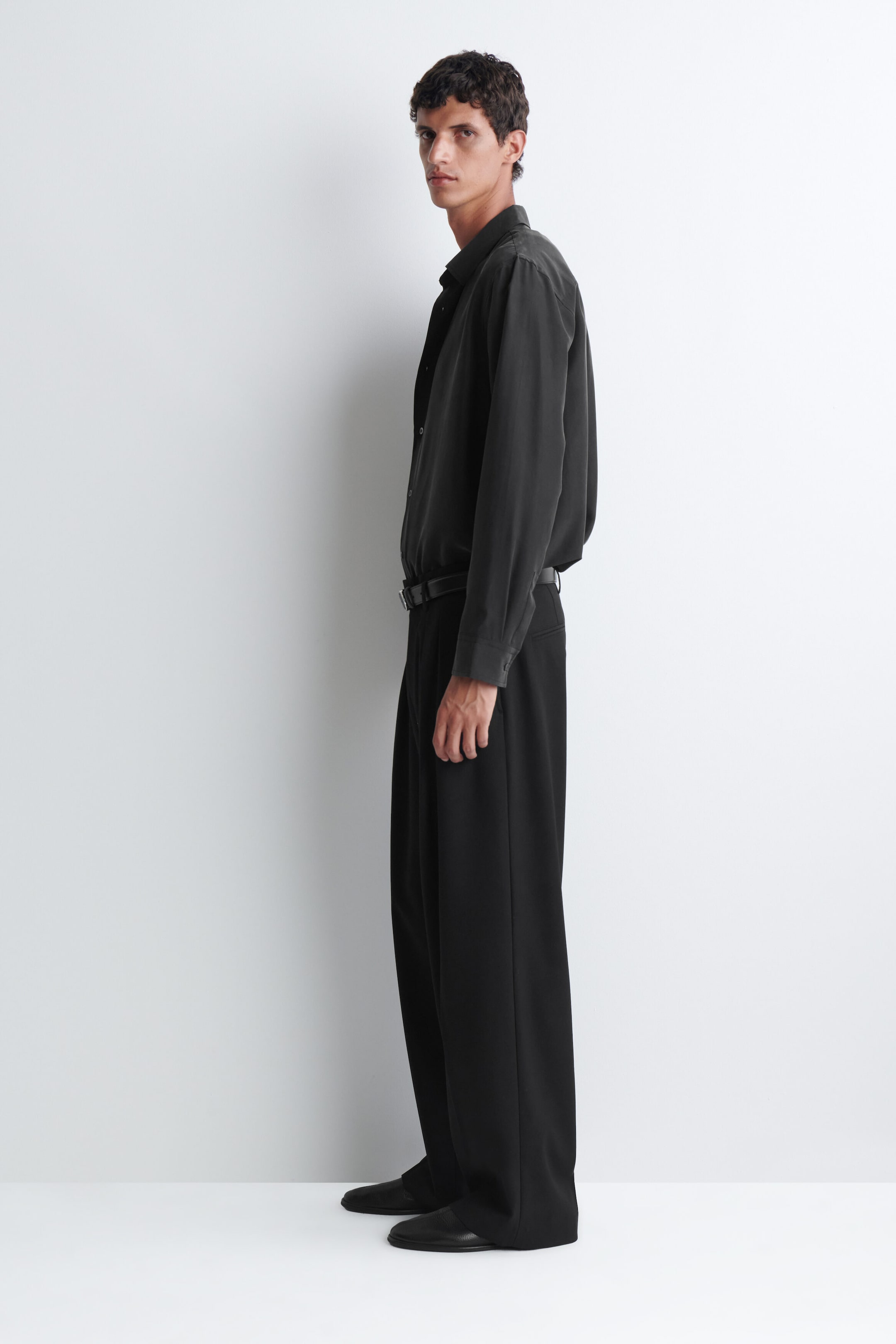 A black sandwashed silk shirt featuring a classic collar and long sleeves is being worn by the model. The shirt has a soft drape with a matte texture and is styled tucked into black wide-leg trousers. A black sandwashed silk shirt featuring a classic collar and long sleeves is being worn by the model. The shirt has a soft drape with a matte texture and is styled tucked into black wide-leg trousers.