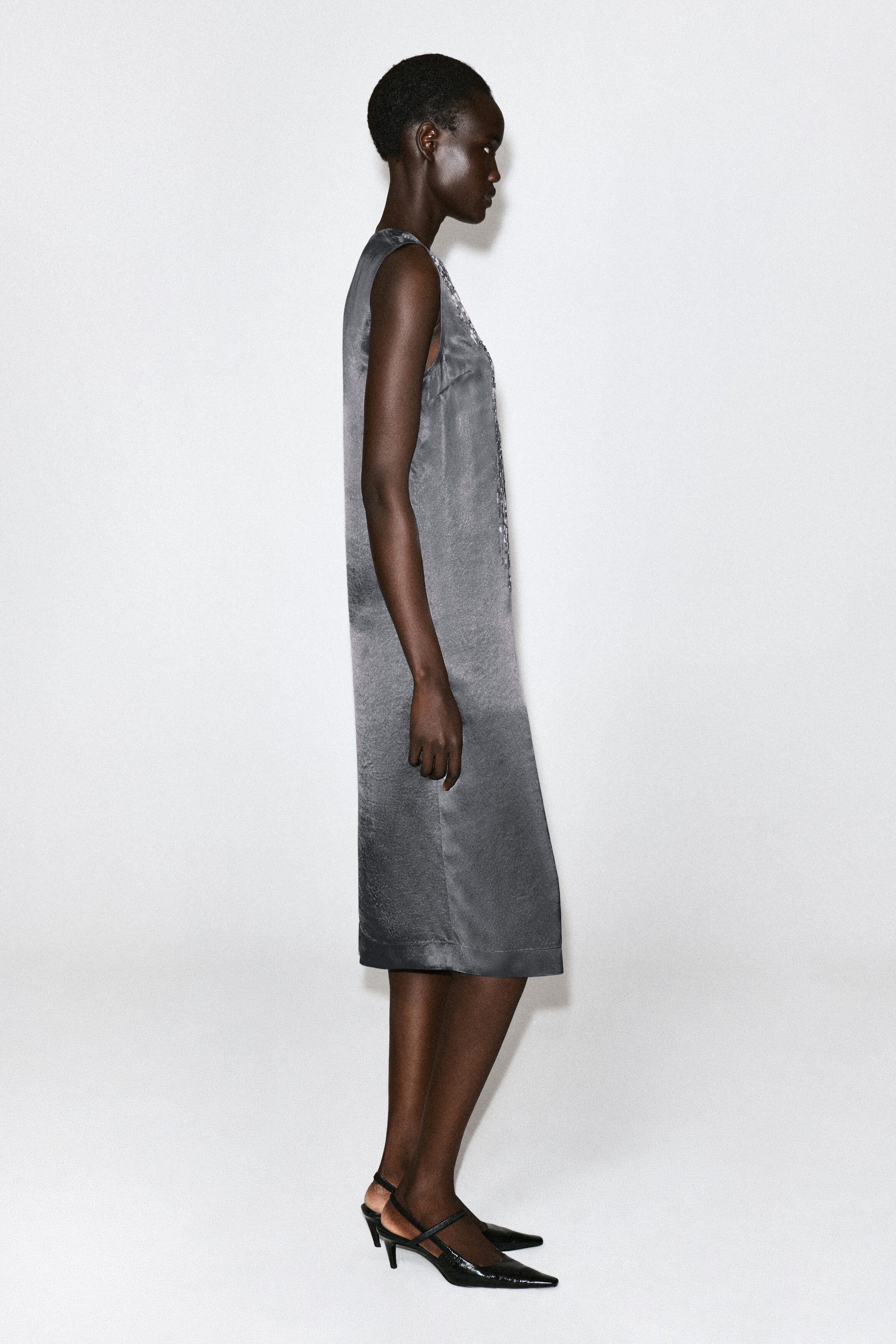 The grey beaded satin V-neck midi dress is being worn and faces left, showcasing its straight, sleeveless silhouette and subtle sheen. Intricate silver beaded embellishments detail the V-neckline, and the dress falls to a midi length. The grey beaded satin V-neck midi dress is being worn and faces left, showcasing its straight, sleeveless silhouette and subtle sheen. Intricate silver beaded embellishments detail the V-neckline, and the dress falls to a midi length.