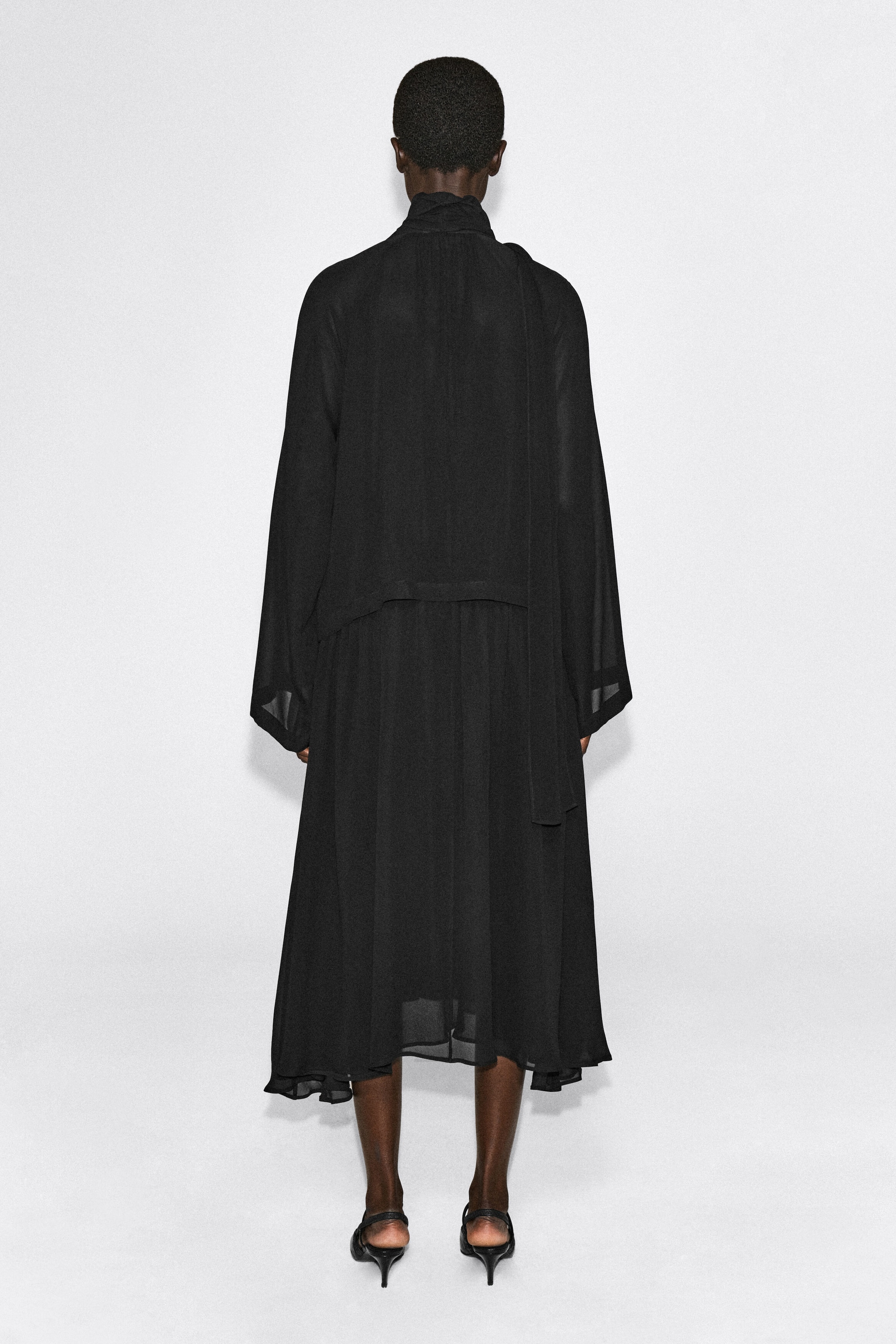 A black fluid midi dress with scarf detail is shown from the back, highlighting its relaxed silhouette and lightweight, semi-sheer fabric. The dress features a high collar with an integrated scarf draping down the side, loose long sleeves, and a layered midi-length skirt.