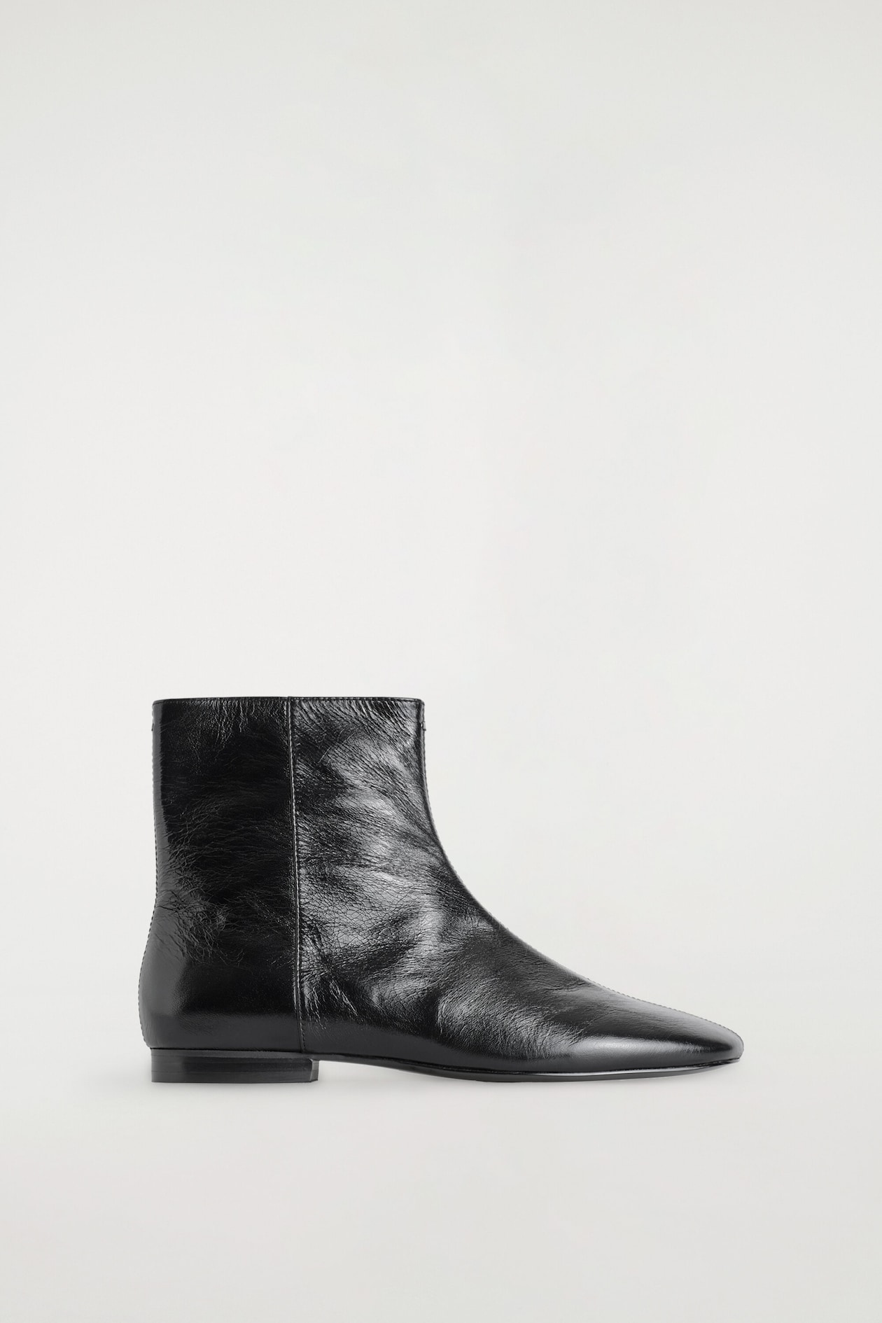 A black crinkled leather ankle boot is facing right, displaying a glossy, textured finish and a minimalist silhouette. The boot features a tapered almond toe, a clean vertical side seam, and a very low, flat block heel.