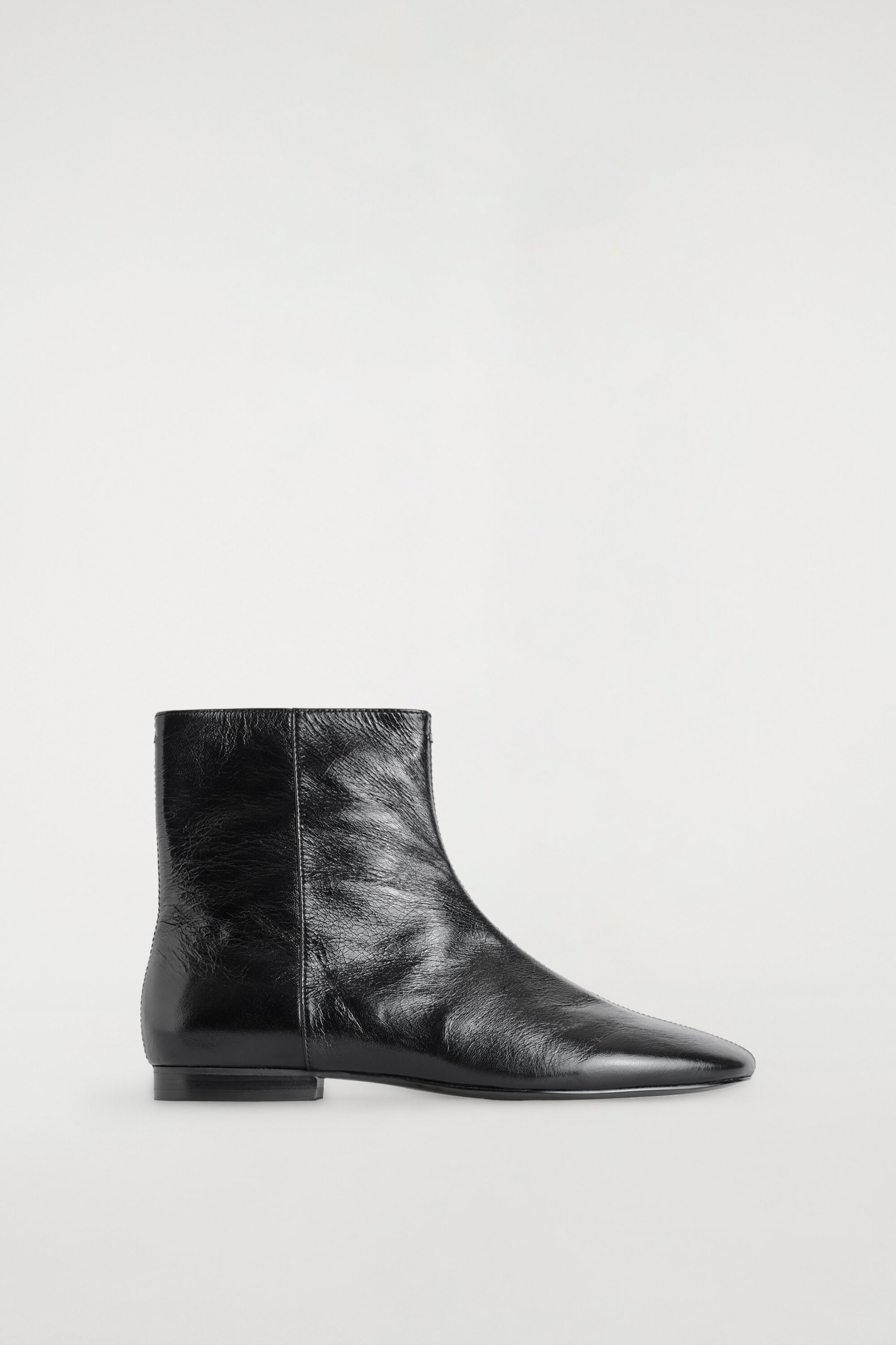 A black crinkled leather ankle boot is facing right, displaying a glossy, textured finish and a minimalist silhouette. The boot features a tapered almond toe, a clean vertical side seam, and a very low, flat block heel.