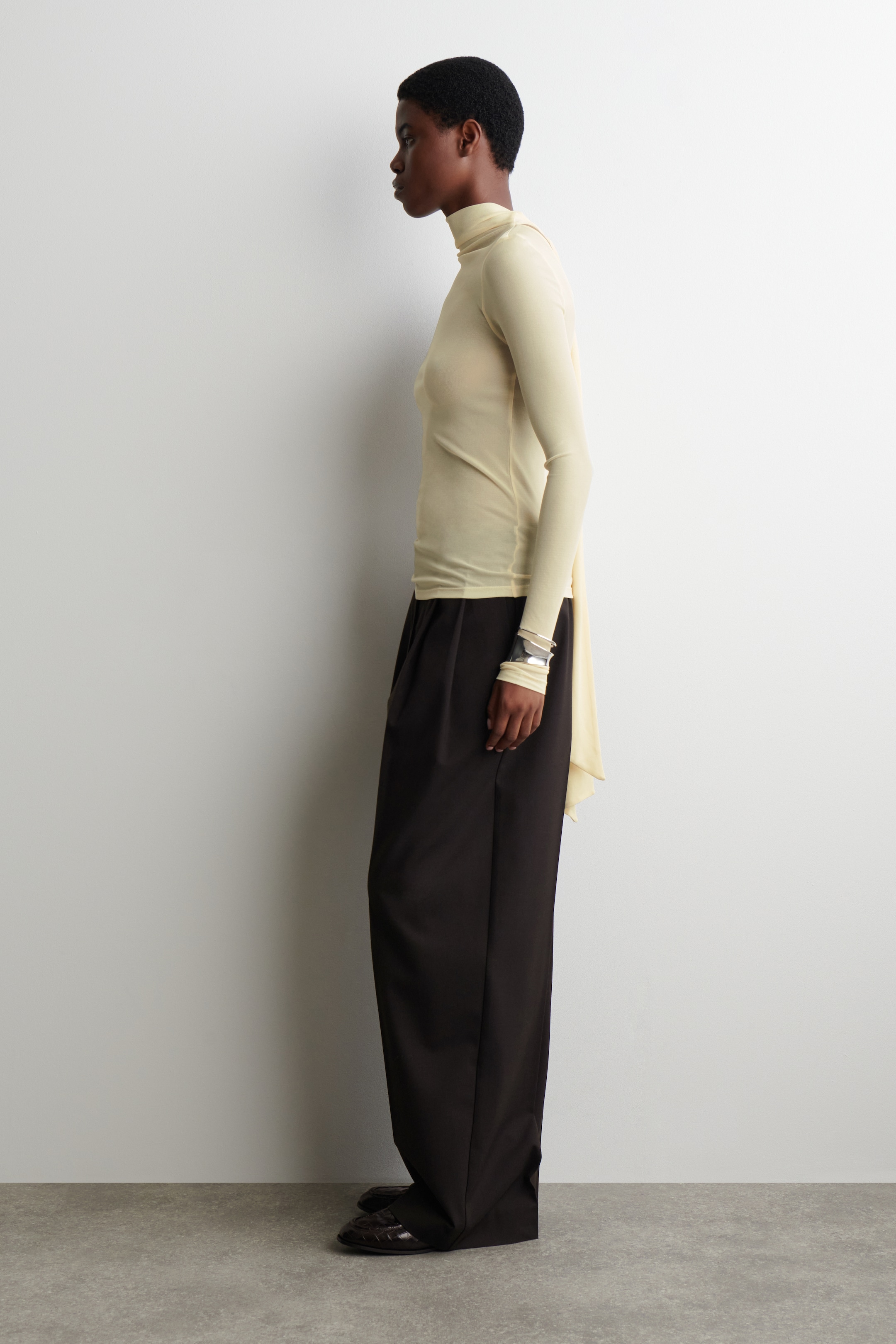 Agrandir l'image: Un haut jaune pâle, semi-transparent, est porté par le mannequin. Le haut présente un col montant et des manches longues, avec un détail d'écharpe ton sur ton qui s'enroule autour du cou et descend le long du dos du mannequin.