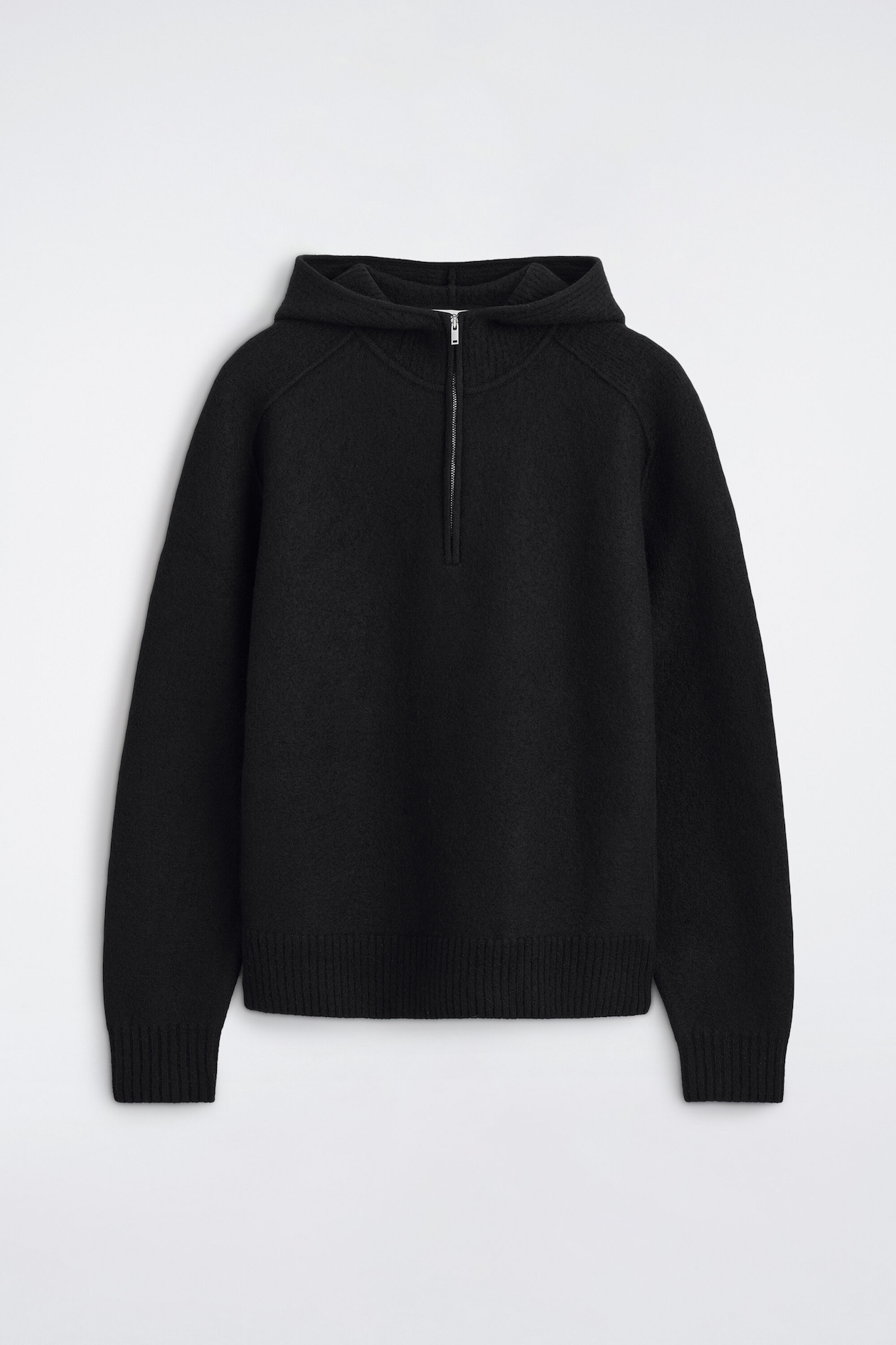 A black merino wool half-zip hoodie is facing the front, showcasing an integrated hood and a silver metal zip fastening. The garment features long raglan sleeves, ribbed cuffs, and a ribbed hem.