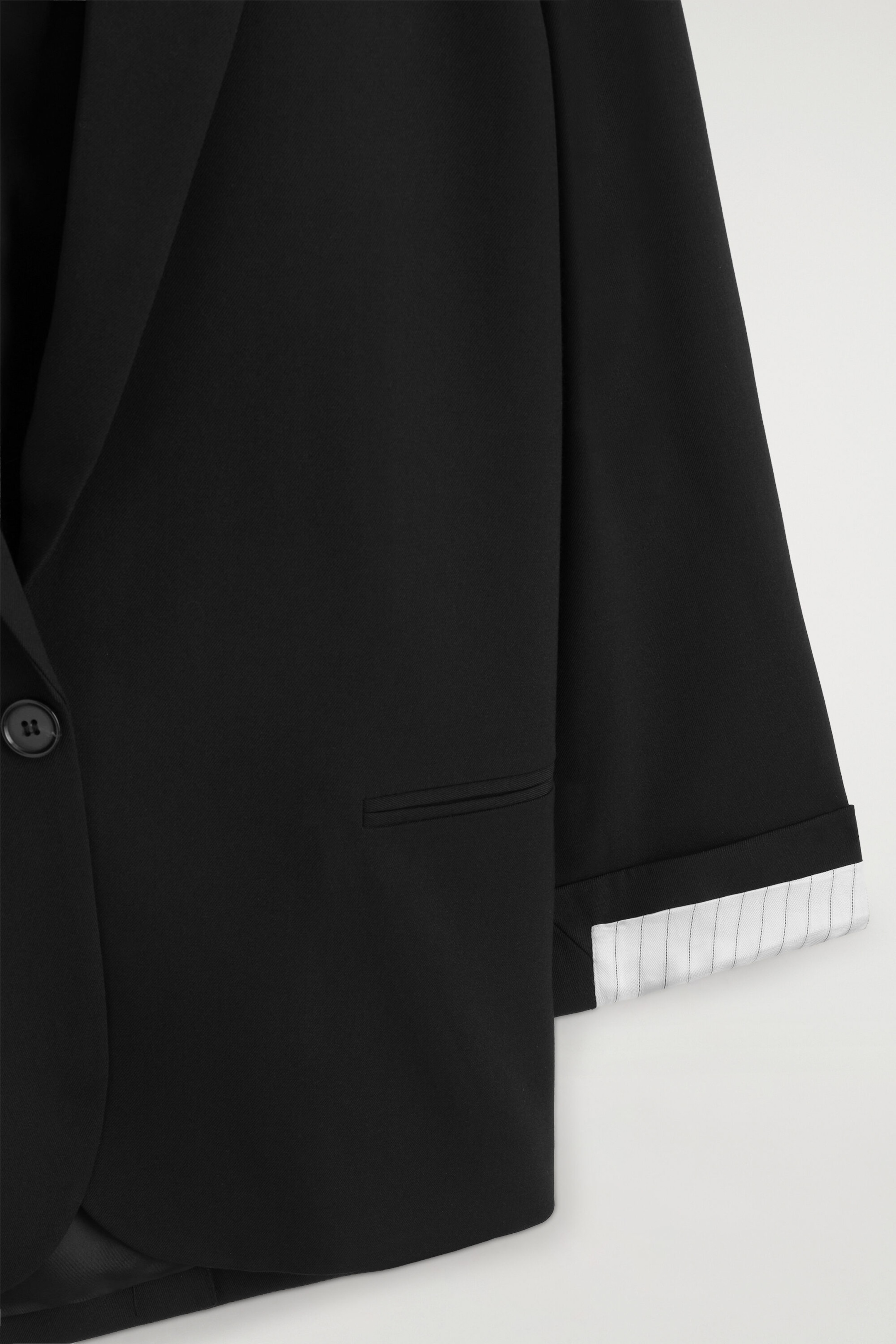 A black blazer is shown in a close-up, front-facing view, featuring a single-button fastening, a notch lapel, and a horizontal jetted pocket. The sleeve is displaying a turned-up cuff, revealing a contrasting white lining patterned with thin, vertical black pinstripes. A black blazer is shown in a close-up, front-facing view, featuring a single-button fastening, a notch lapel, and a horizontal jetted pocket. The sleeve is displaying a turned-up cuff, revealing a contrasting white lining patterned with thin, vertical black pinstripes.