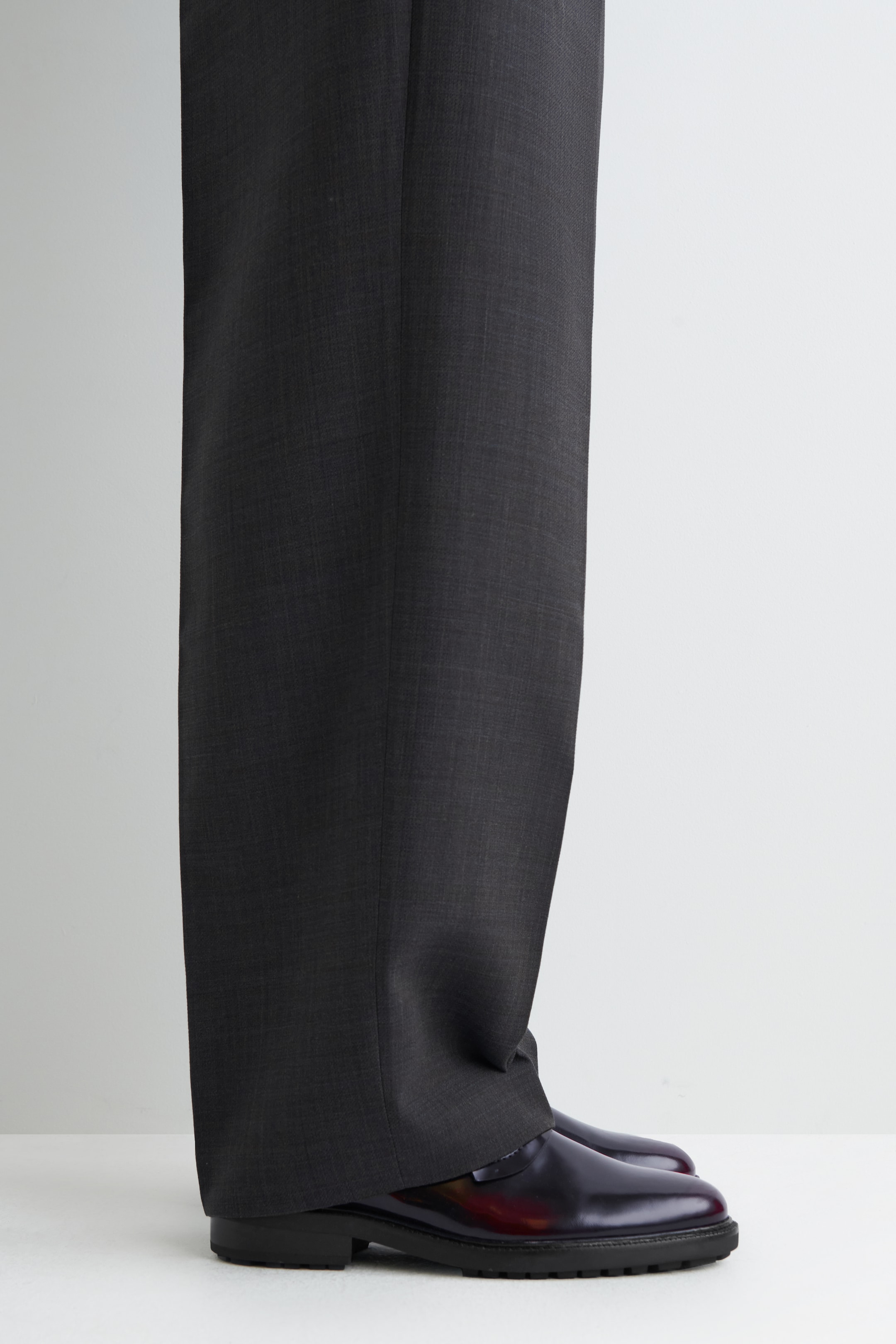 Ingrandisci l'immagine: Un paio di pantaloni a gamba larga in carbone scuro si drappeggia su mocassini bordeaux lucidi, mostrati da un'angolazione laterale. I pantaloni presentano una trama sottile e testurizzata, e i mocassini sfoggiano una spessa suola nera a carrarmato.