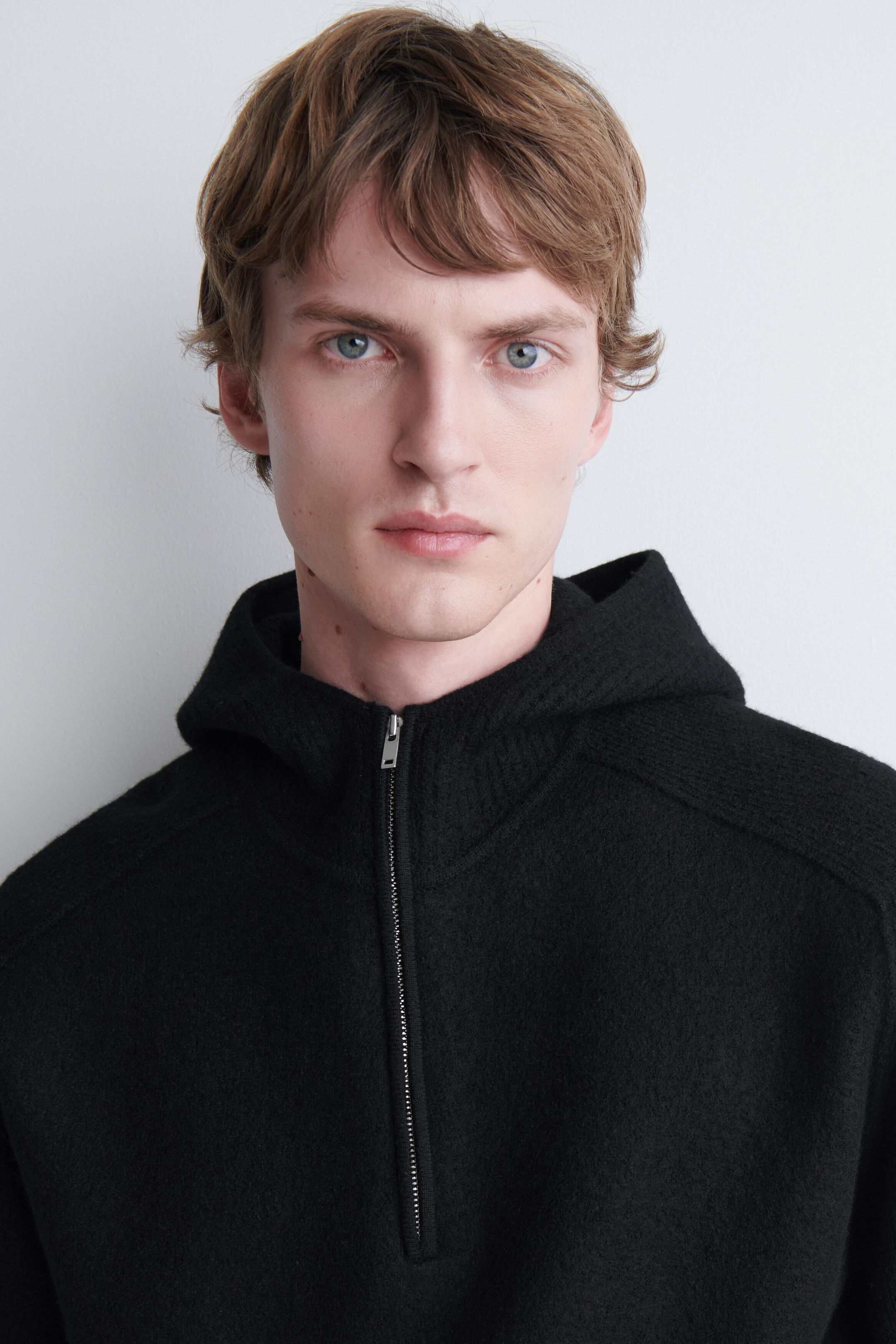 A black merino wool half-zip hoodie featuring a textured knit, an attached hood, and a silver zip is being worn by the model. The zip is partially open at the high collar. A black merino wool half-zip hoodie featuring a textured knit, an attached hood, and a silver zip is being worn by the model. The zip is partially open at the high collar.