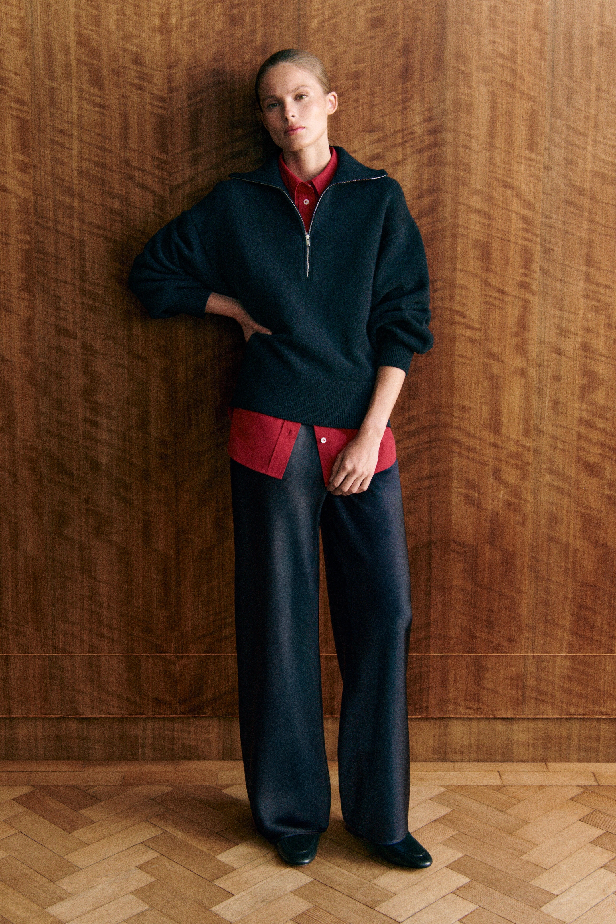 A dark navy knitted sweater is shown from the front, featuring a relaxed fit, sculptural sleeves, and a half-zip funnel neckline with a silver-tone zipper. The sweater is layered over a bright red collared shirt, with the zipper partially unzipped to reveal the collar. A dark navy knitted sweater is shown from the front, featuring a relaxed fit, sculptural sleeves, and a half-zip funnel neckline with a silver-tone zipper. The sweater is layered over a bright red collared shirt, with the zipper partially unzipped to reveal the collar.